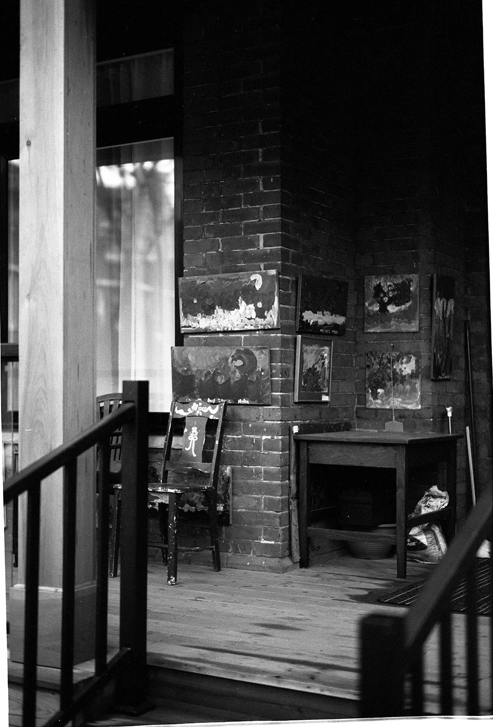 A corner of an art studio with paintings on brick and wood walls, a painted chair, and a table with art supplies, in black and white.