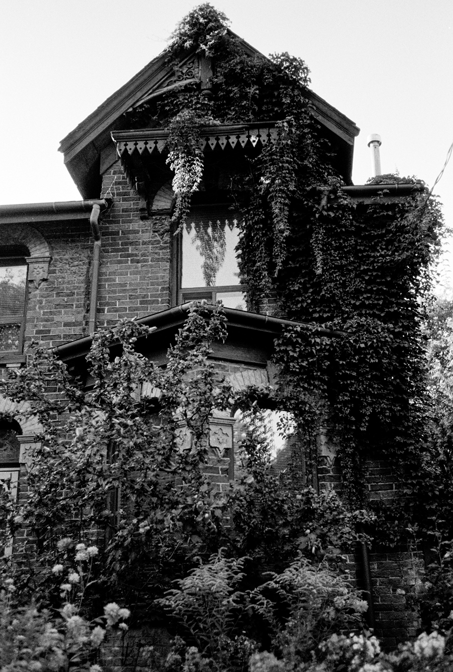 Black and white photo of an old brick house with ivy growing on the walls and around the windows, featuring a gabled roof and decorative trim.
