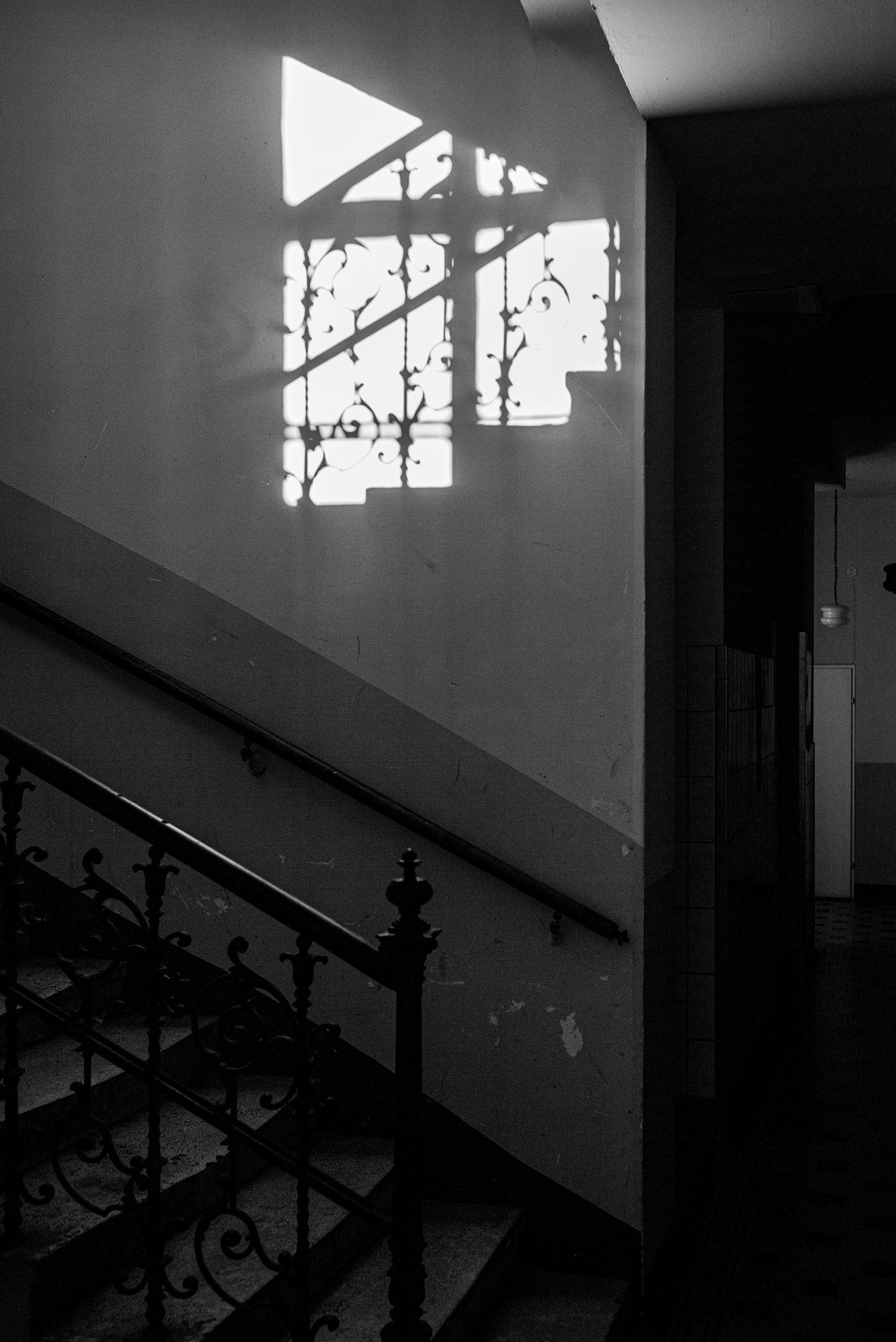 Sunlight creates a shadow of an ornate window grill on a white wall inside a building, with a staircase with decorative railing in the foreground.