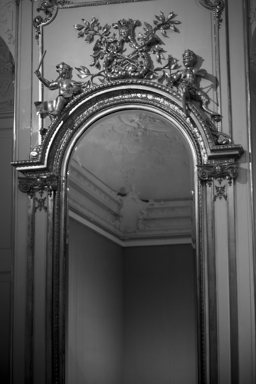 An ornate mirror with decorative carvings and figurines of children on top, set in a classical or baroque styled room.
