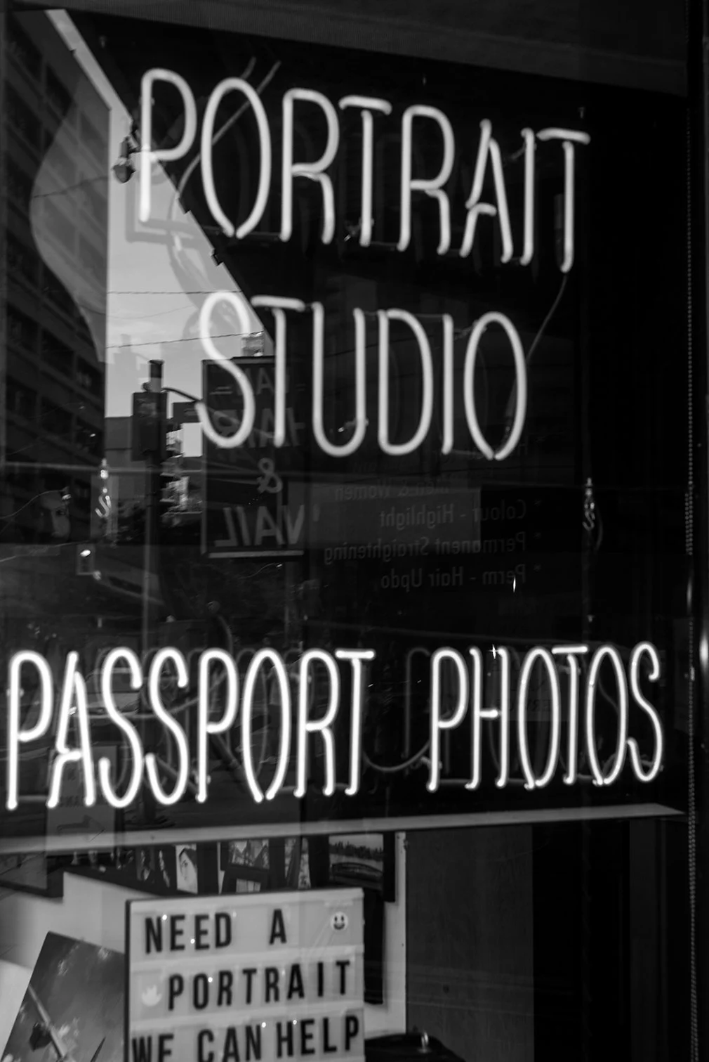 Neon sign reading 'Portrait Studio & Passport Photos' in black and white, with reflections of city buildings and a smaller sign below saying 'Need a Portrait? We Can Help'.