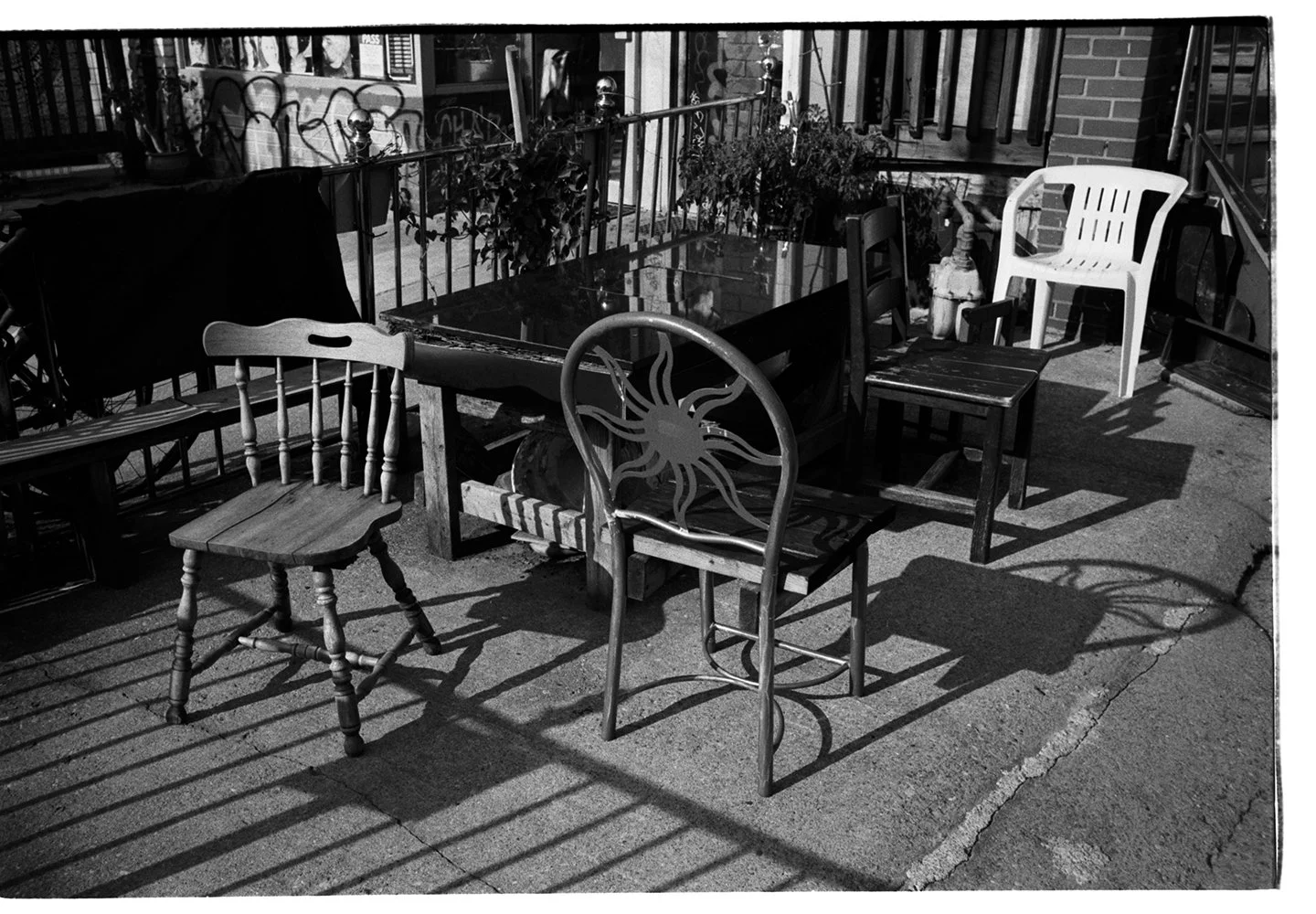 Empty outdoor patio with various chairs surrounding a glass table, along with potted plants and graffiti on the wall in black and white.