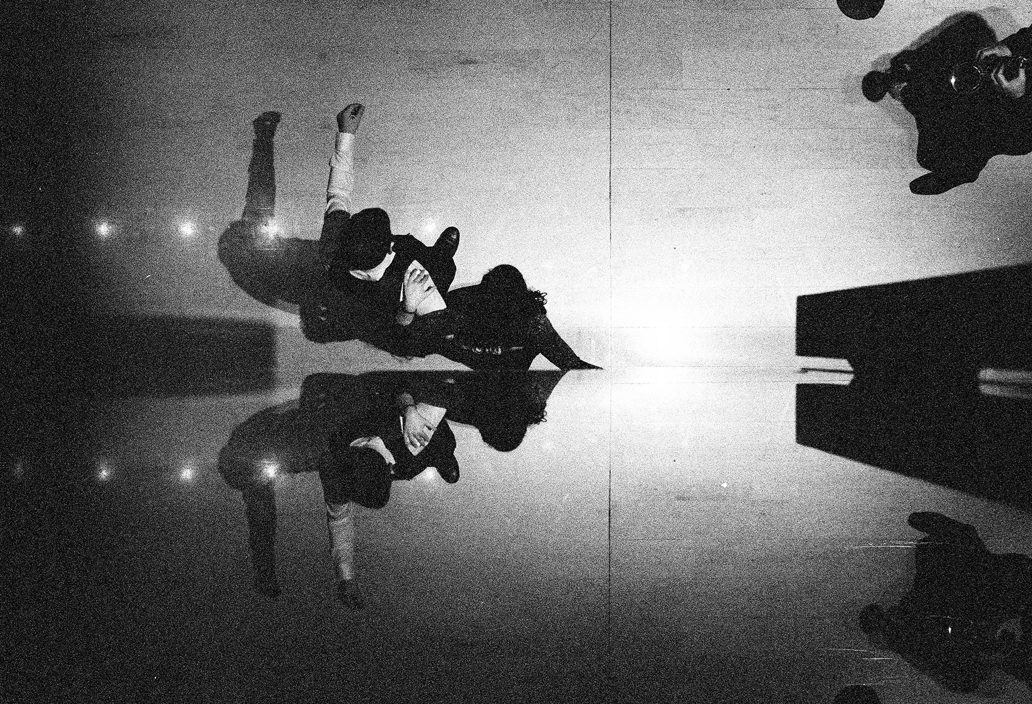 Black and white photo of three people, two women and one man, sitting on a reflective floor, making peace signs with their hands. The scene is indoors, with a plain wall and some lights in the background. The photo has a grainy quality.