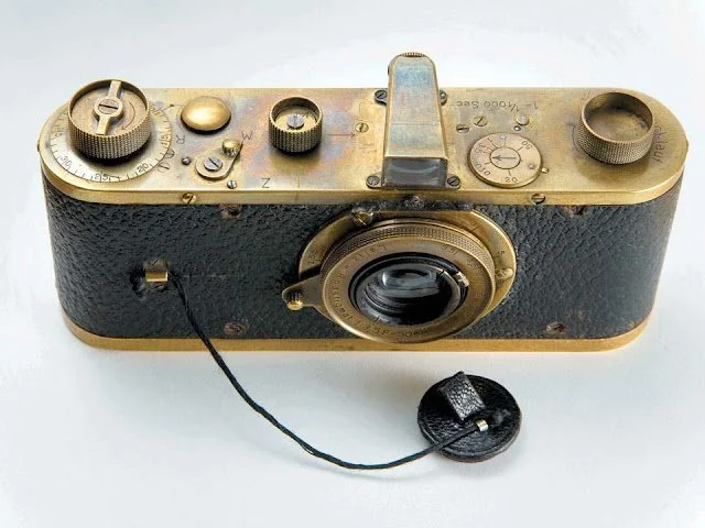 A vintage black and gold camera with a viewfinder, adjustable dials, and a lens, connected to a black strap.