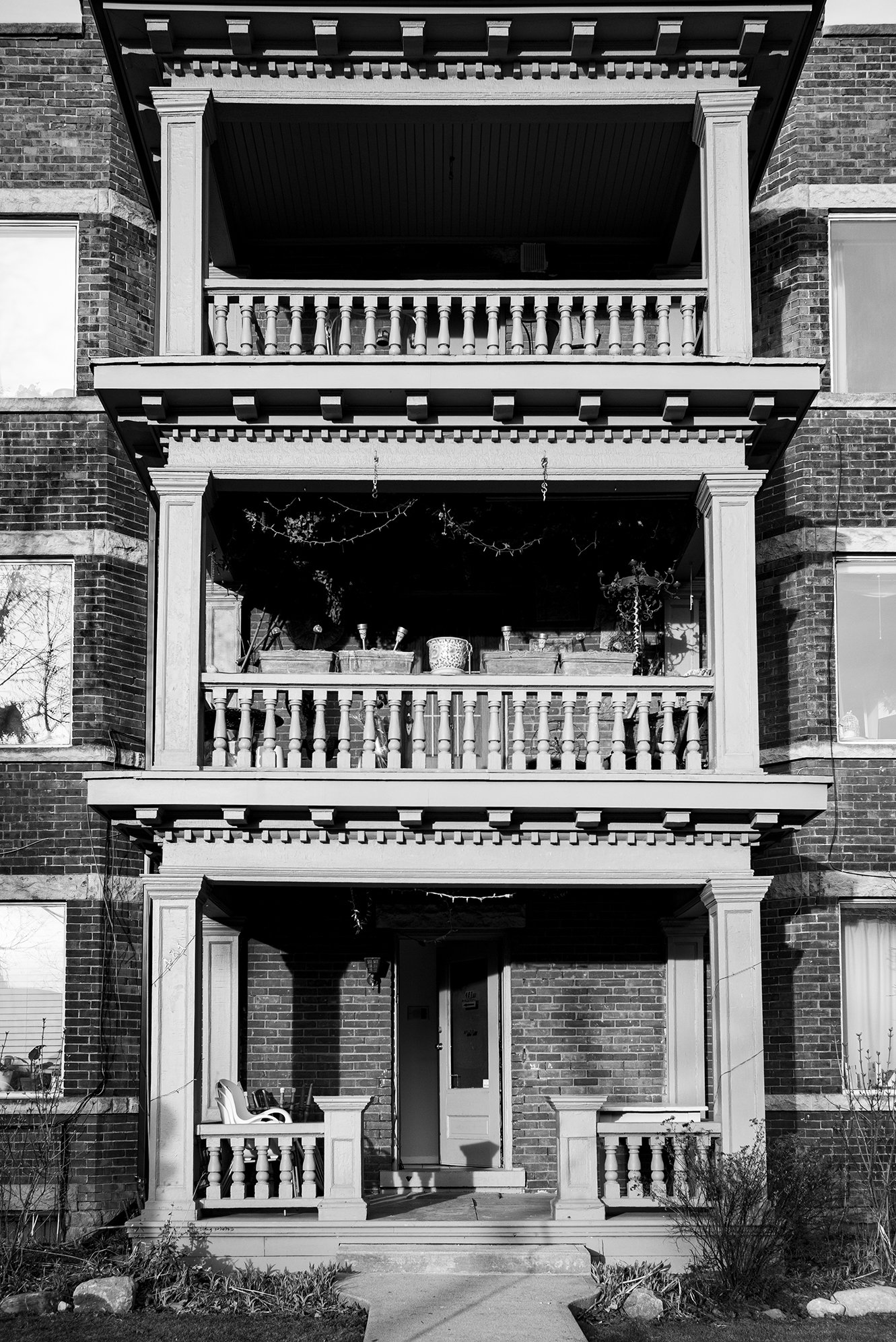 Black and white photo of a three-story brick apartment building with a front porch on each level. The porches have decorative railings and columns. The bottom porch area has a door in the center with chairs and plants on the porch.