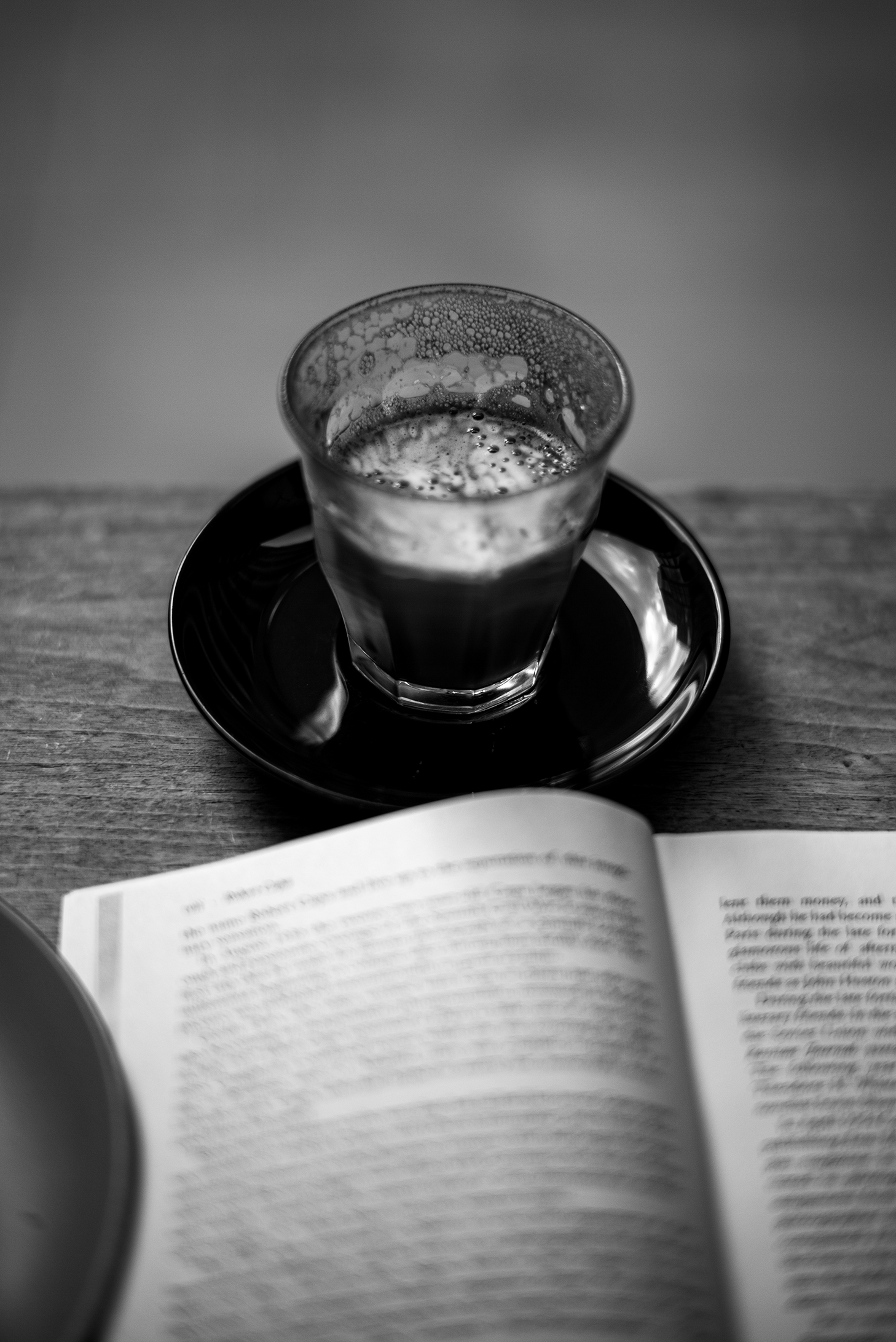 A glass of coffee on a saucer next to an open book on a wooden table, in black and white.