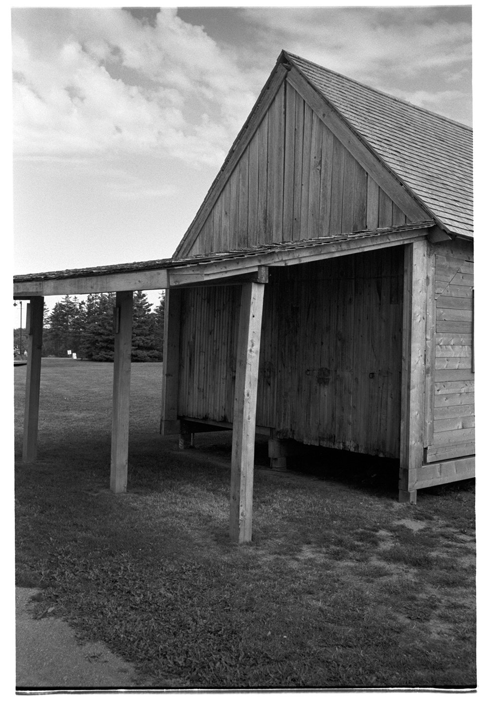 Black and white photo of a wooden barn with a gabled roof, supported by wooden posts, and surrounded by grass and trees in the background.