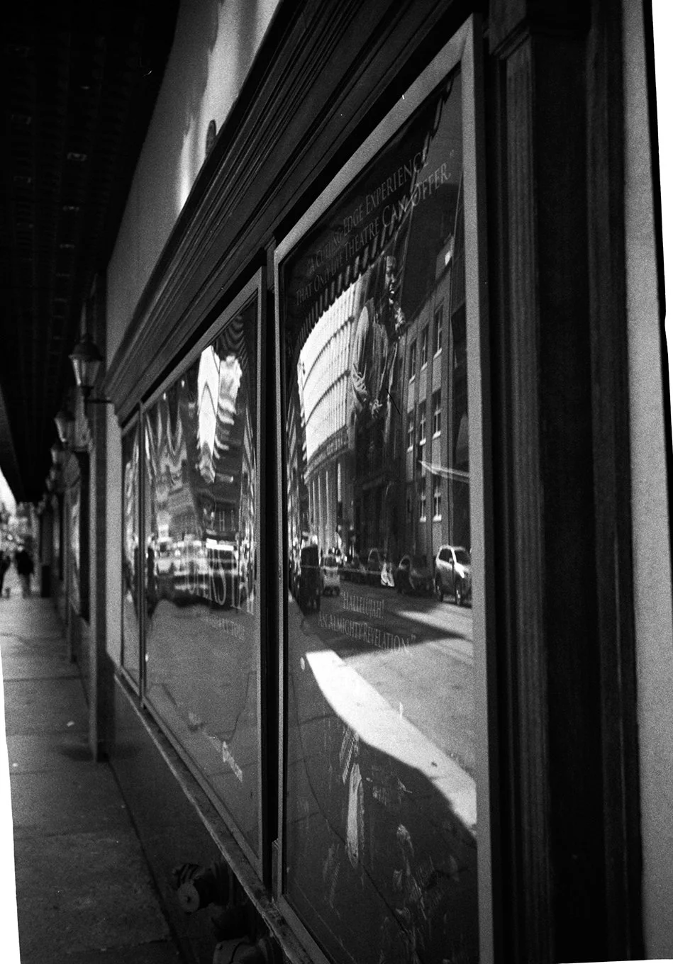 Black and white photo of a row of window display posters on a city sidewalk, with reflections of buildings, cars, and a person visible on the glass.