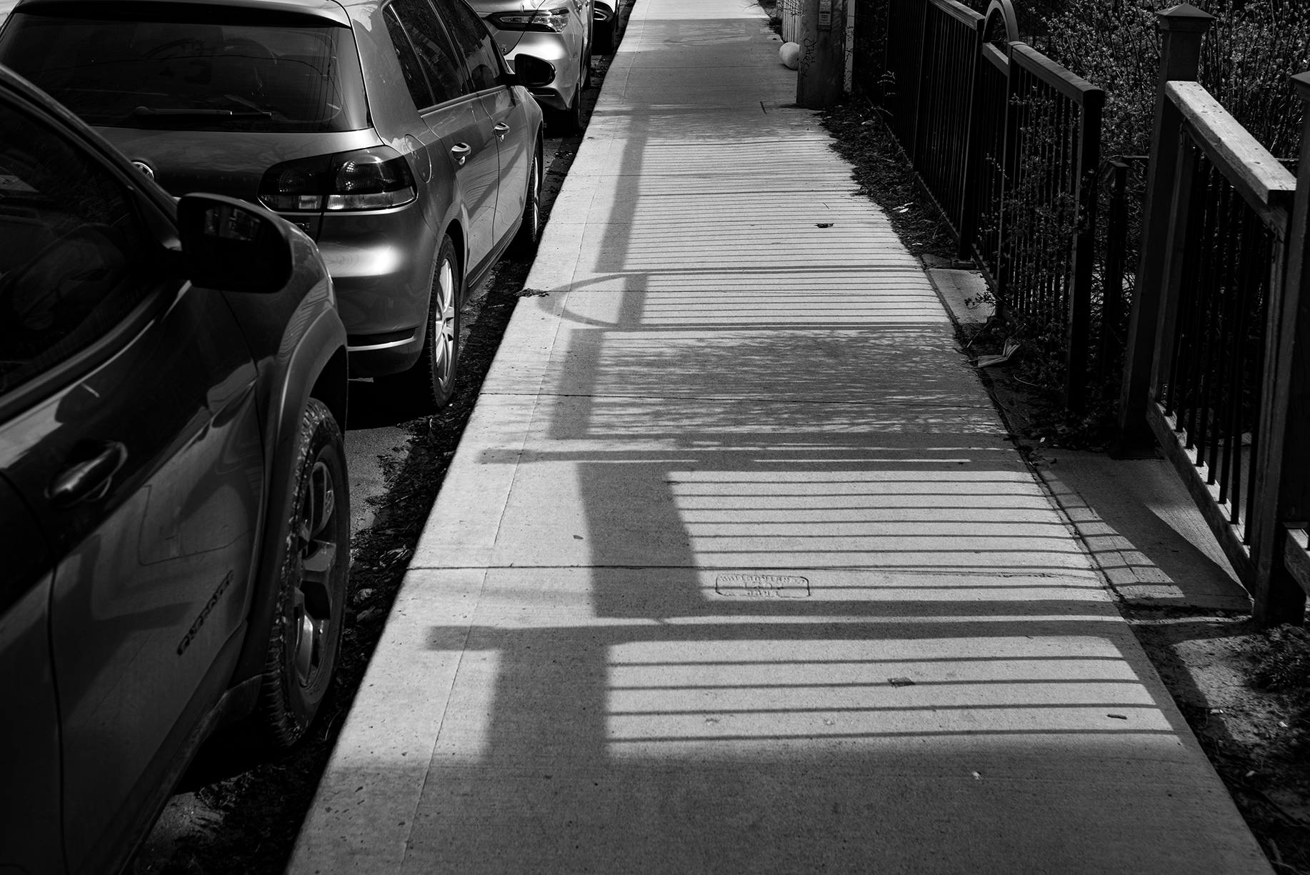 Black and white photo of a sidewalk with shadows of a fence and lamp post, parked cars on the left and a fence on the right side.