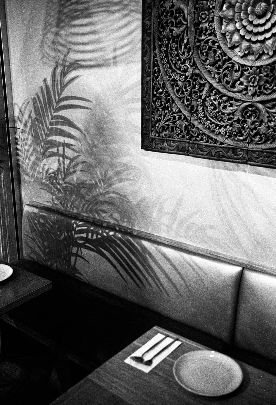 Black and white photo of a restaurant interior with a shadow of a leafy plant cast on the wall, a decorative wooden panel, a padded bench, and a place setting on the table with a plate and cutlery.