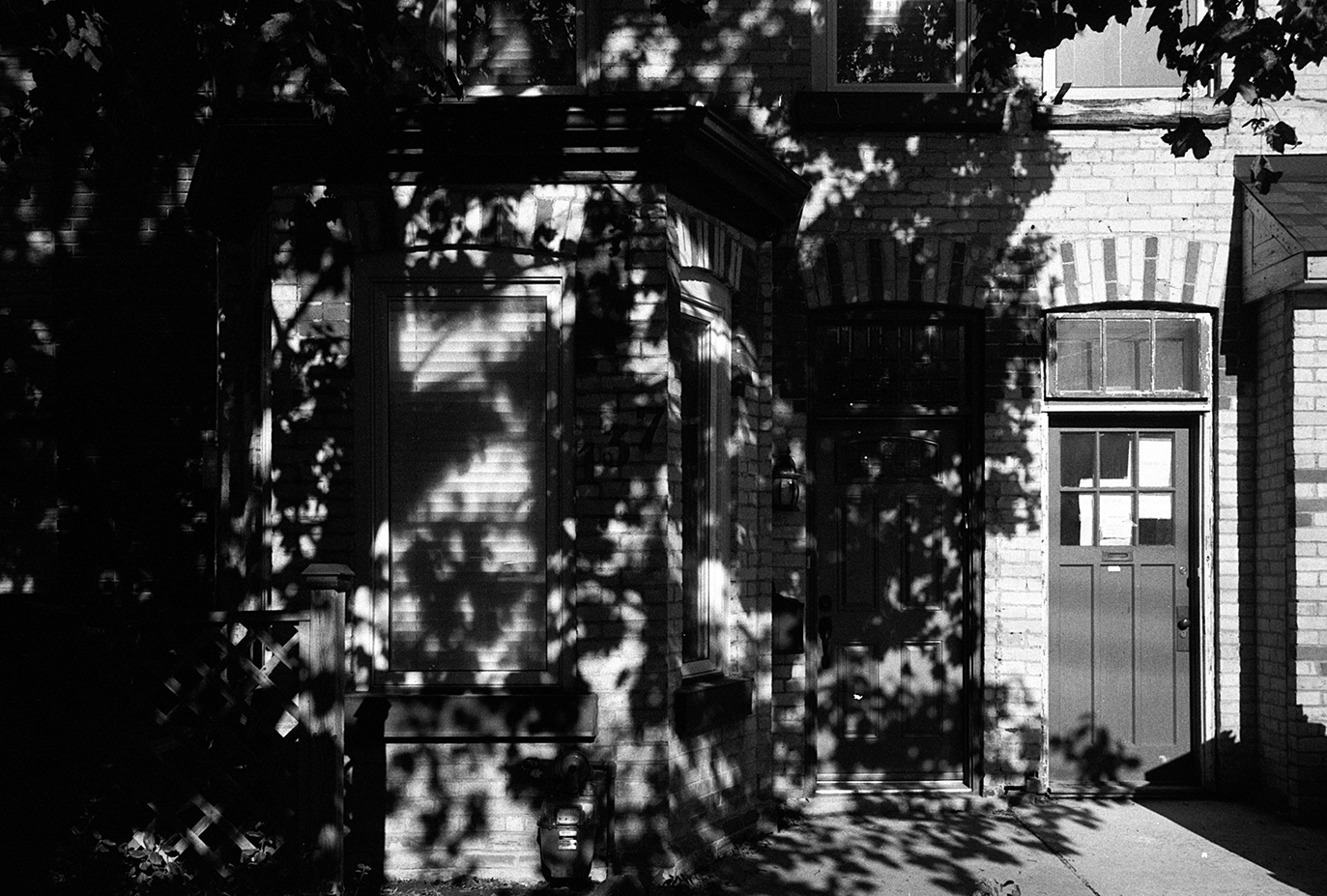 Black and white photo of a brick house front with a door, window, doorbell, and trees casting shadows on the facade.