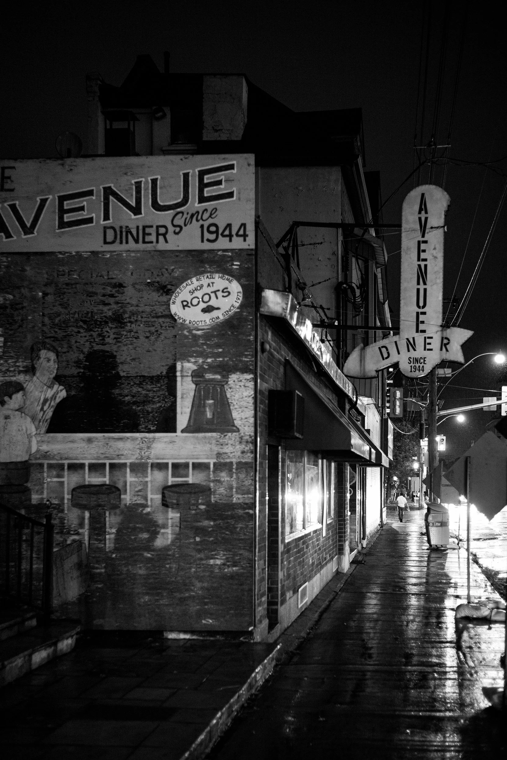 Nighttime street scene featuring a vintage diner with neon illuminated signs reading "Avenue Diner" since 1944. The sidewalk is wet, reflecting nearby lights, and a person is walking in the distance.