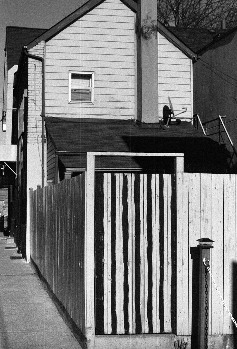 Black and white photo of a residential alleyway with a wooden fence, house, and stairs. A person is standing on the roof holding a broom. There is a small window on the house.
