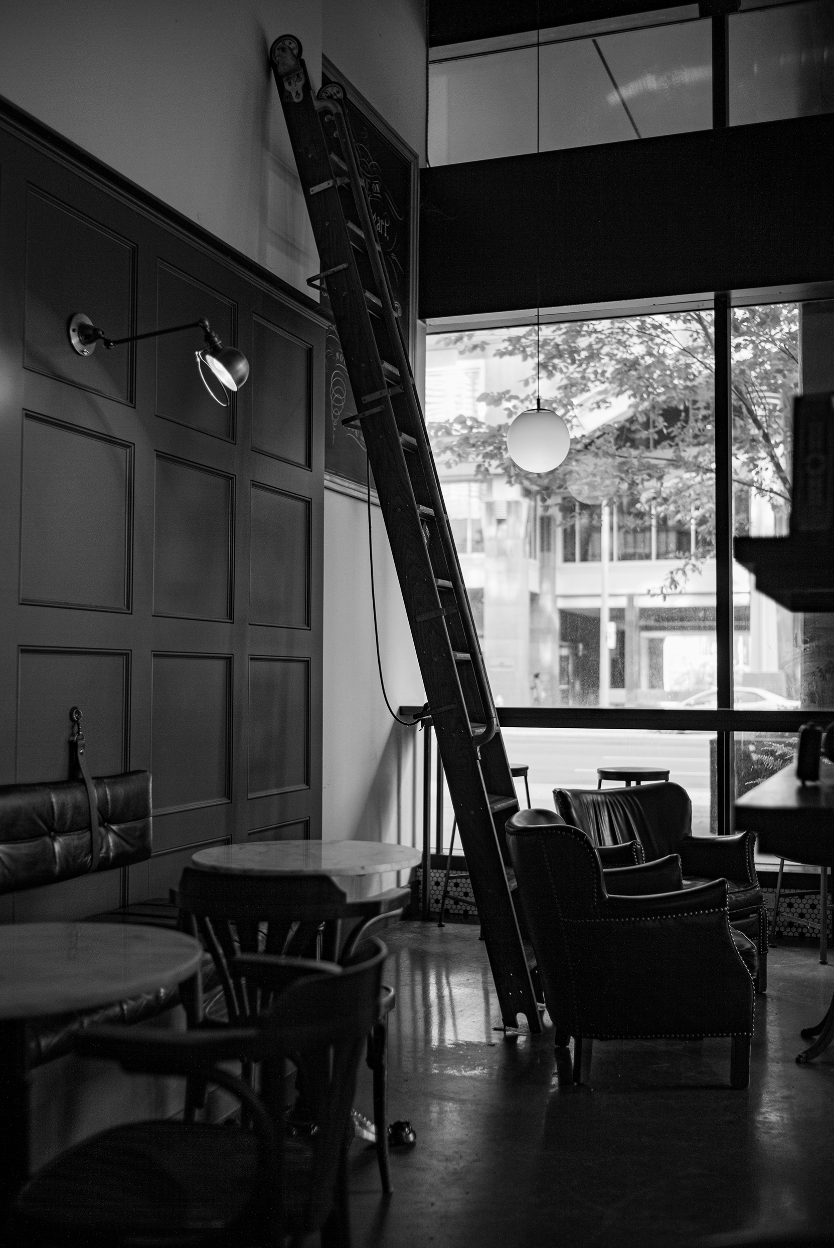 Interior of a cozy cafe with a large window, two leather chairs, a round table, and a ladder leaning against the wall.