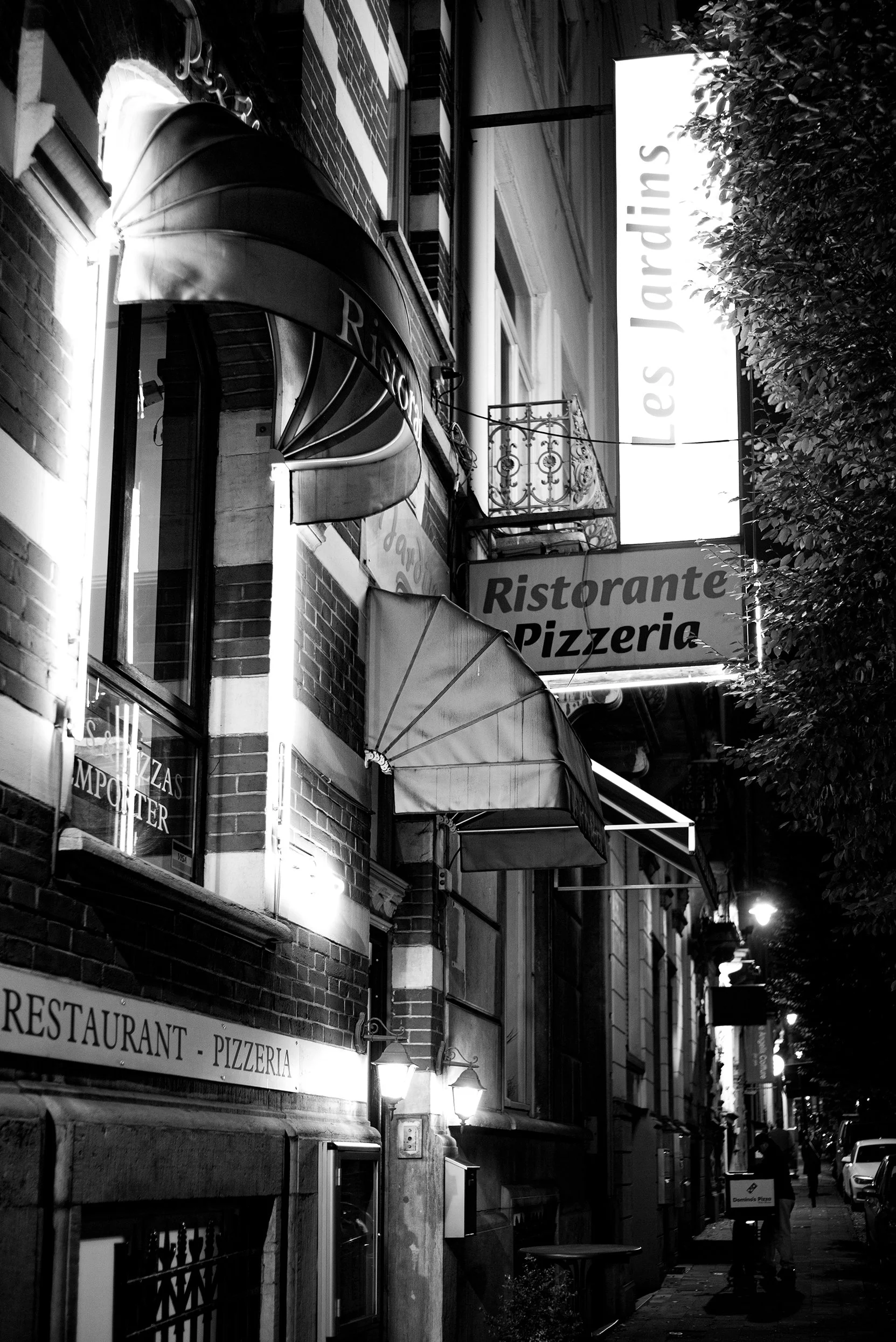 Night street scene with signs for a restaurant and pizzeria, illuminated lamps, and outdoor seating, with a pedestrian walking on the sidewalk.