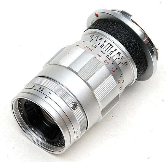 Silver camera lens with various focusing and aperture markings.