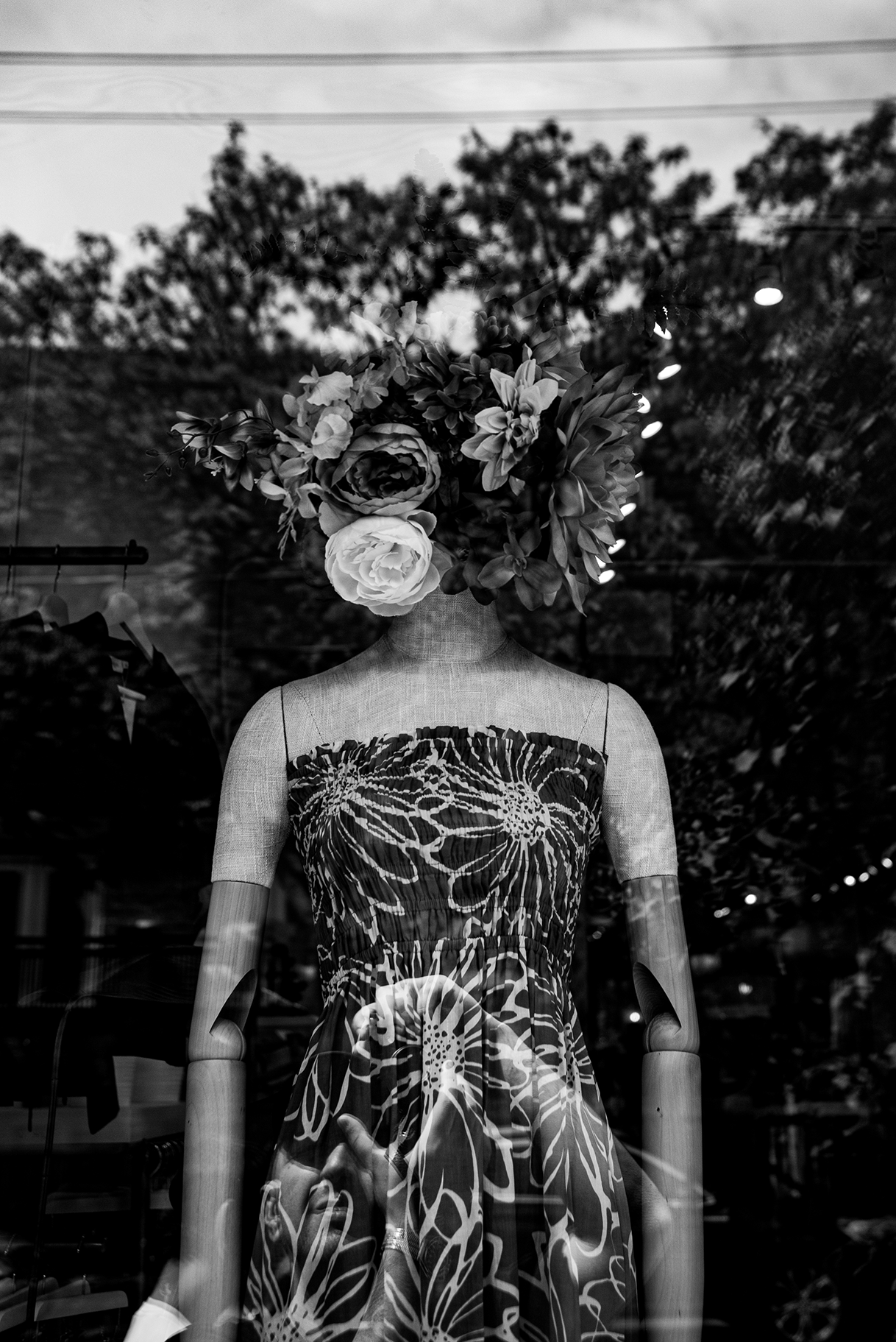 Black and white photo of a mannequin in a store window wearing a dress with a floral pattern and shoulder straps, with a large bouquet of flowers as the mannequin's head.