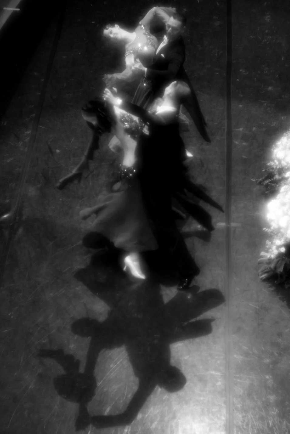 A couple is dancing closely on a reflective floor, viewed from above, with reflections creating a mirror image of their movements.