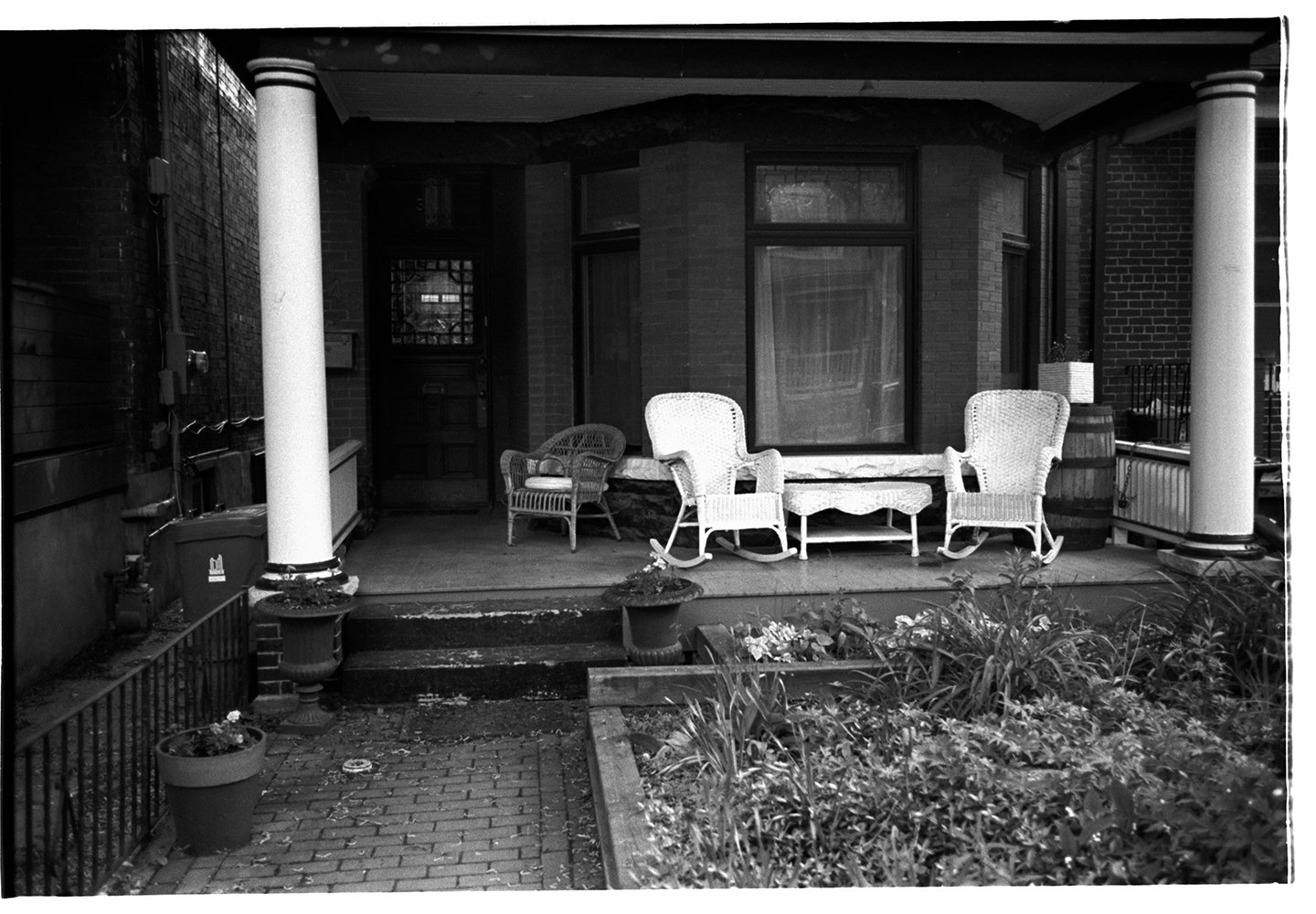 Black and white photo of a porch with wicker chairs, a table, potted plants, and steps leading up to a house with large windows and brick walls.