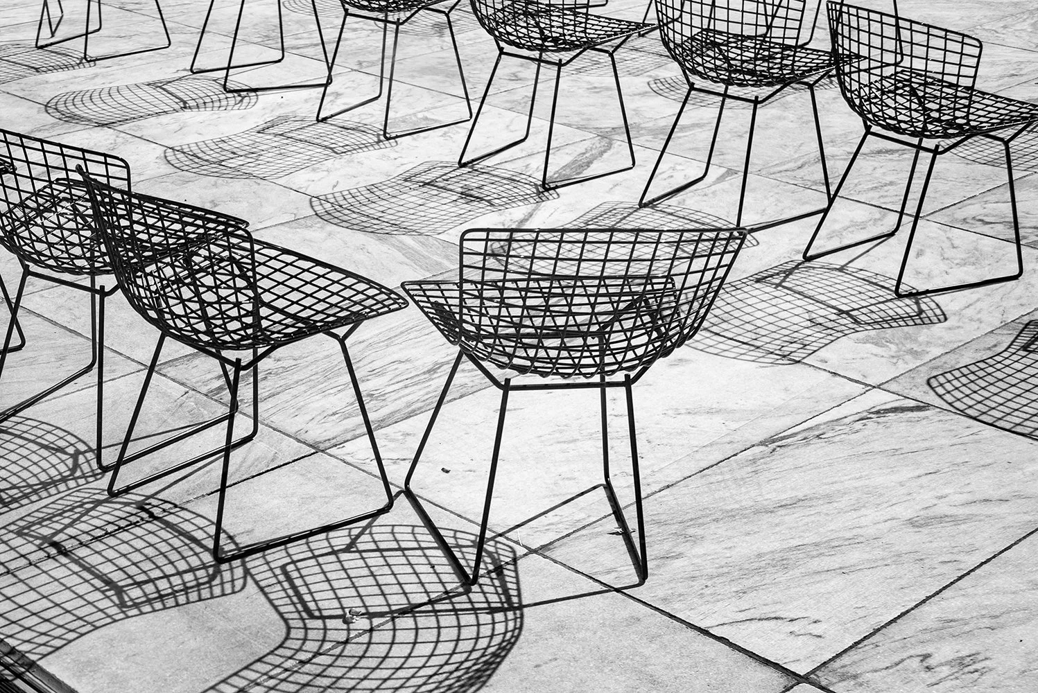 Black wireframe chairs arranged on a tiled floor, casting shadows.
