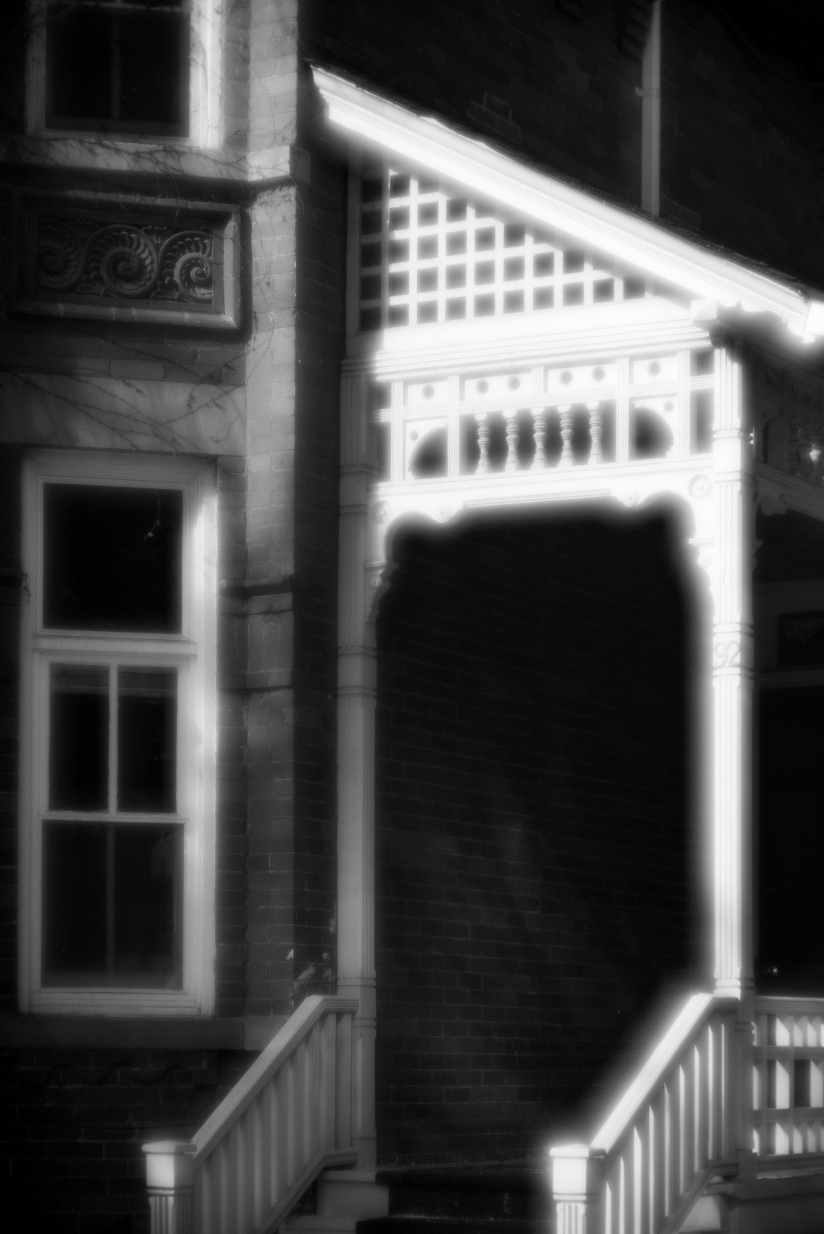 Black and white photo of Victorian-style house entrance with stairs, porch, decorative railing, and window, illuminated by bright light.