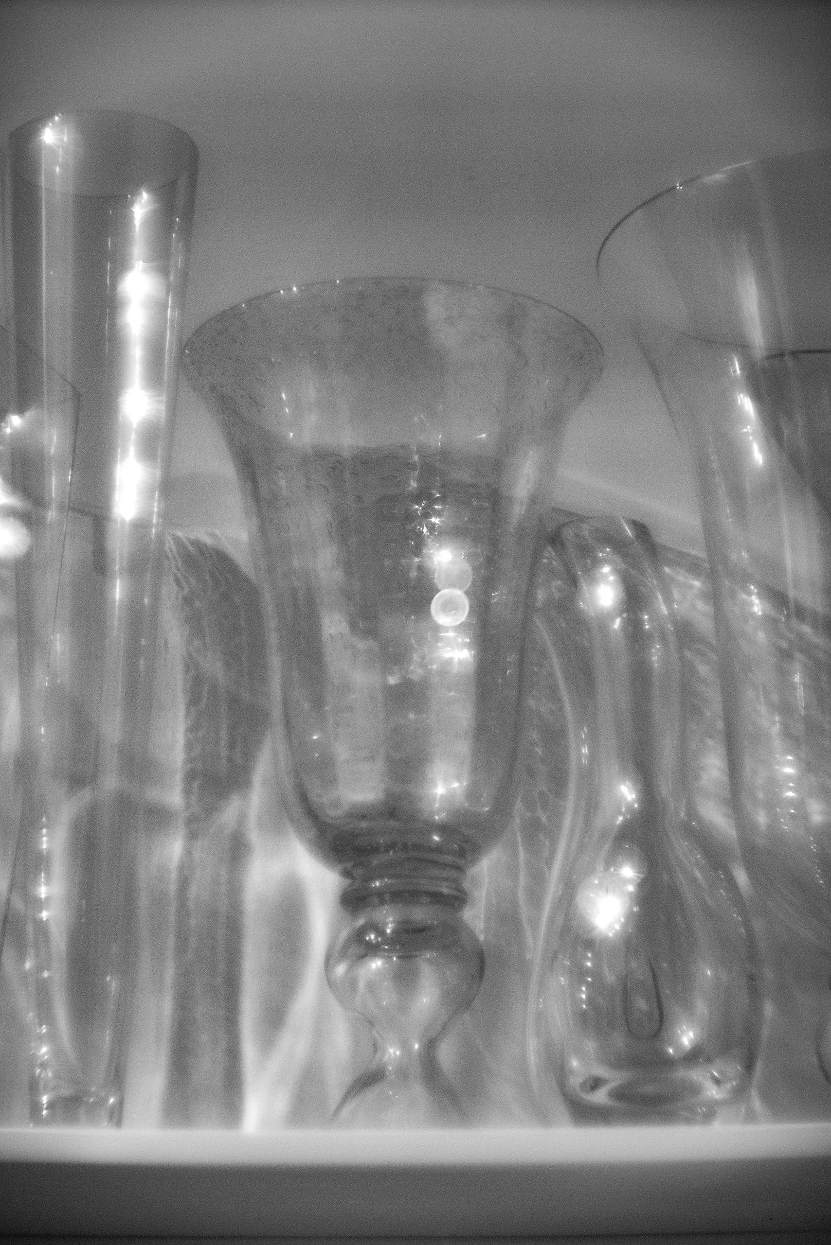 Several glass vases and containers arranged together, including a tall cylindrical vase, a large experimental vase, and a round-bottomed vase, all made of clear glass, with reflections and light bouncing off their surfaces.