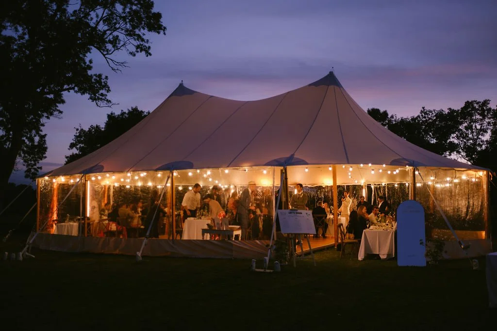 A large outdoor event tent illuminated with string lights during dusk, with people dining inside at tables.