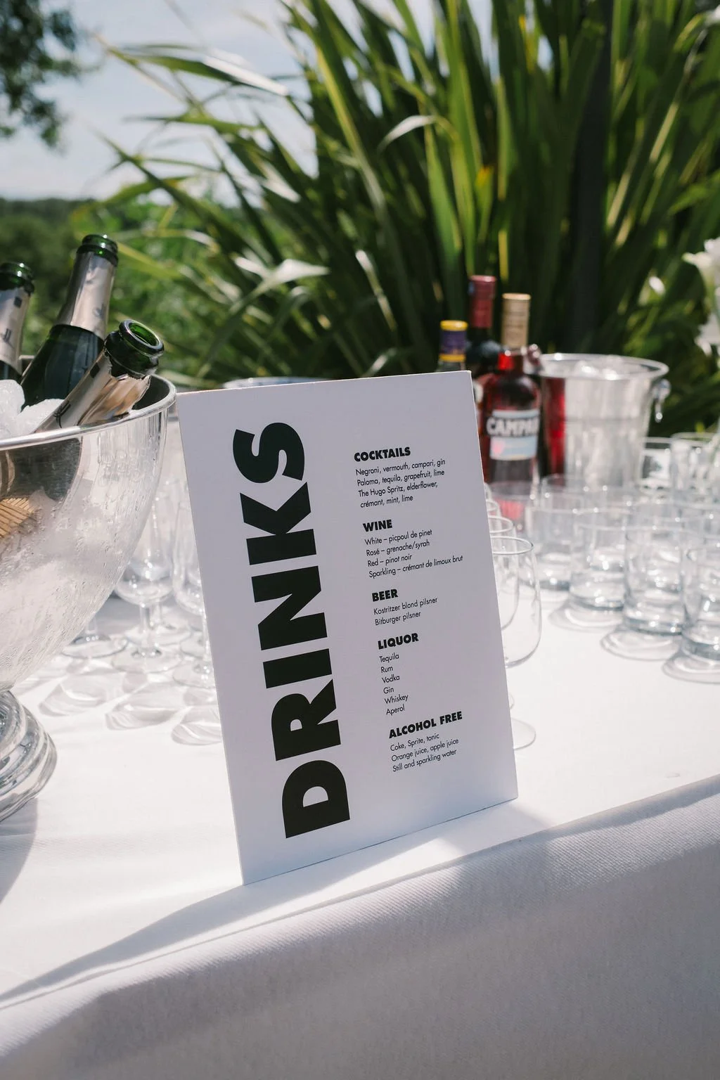 A drink menu on a white table outside with bottles of alcohol and ice buckets.