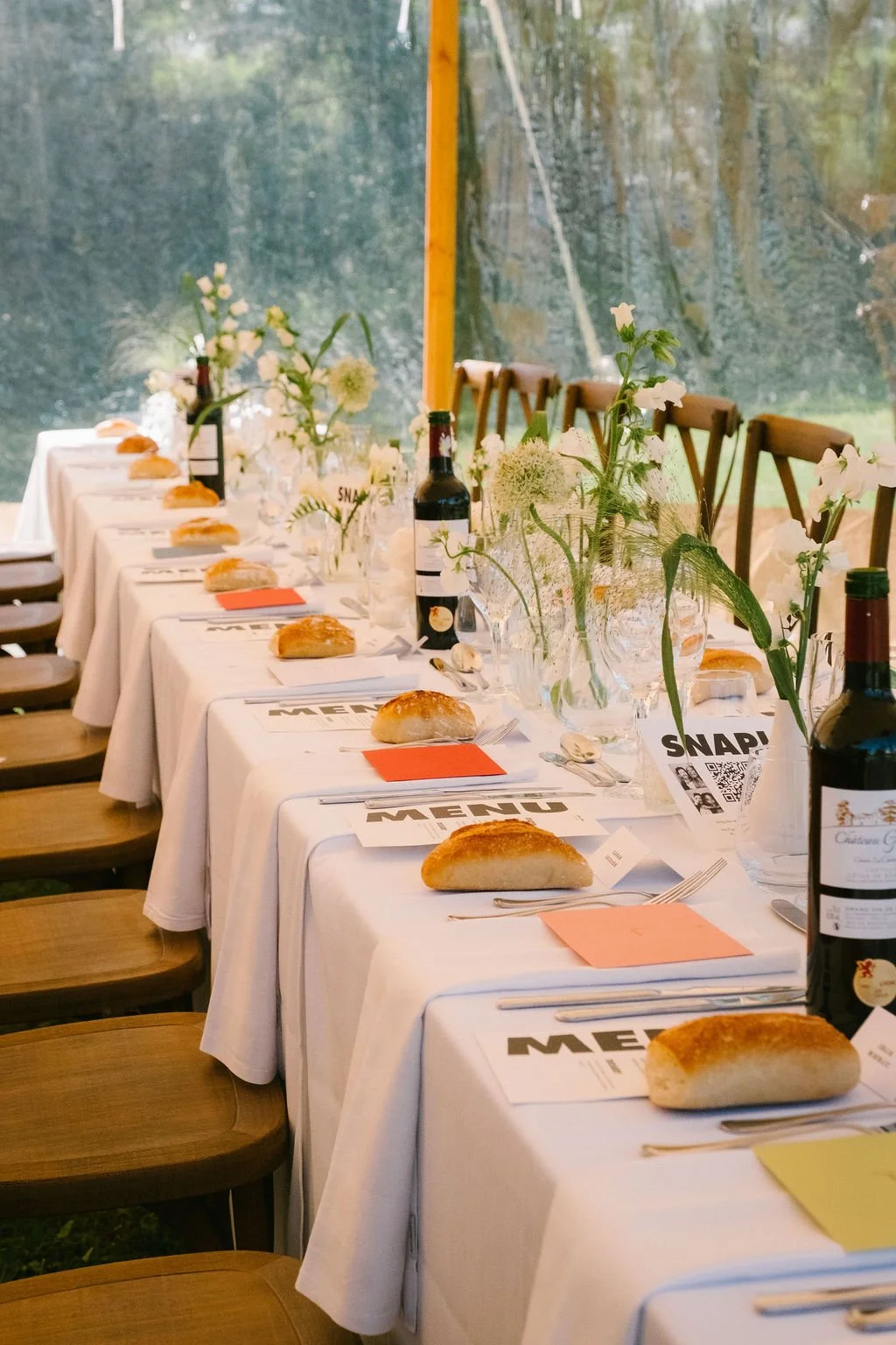 A long dining table set for a meal outside with white tablecloths, floral centerpieces and menus.