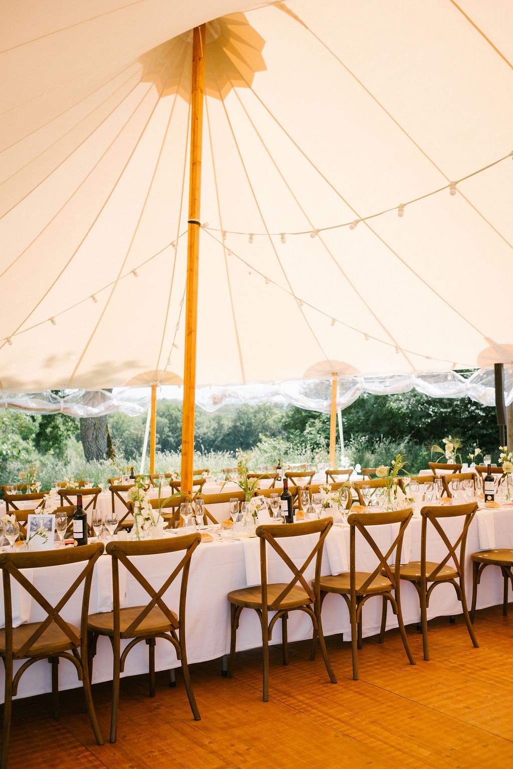 An outdoor dining setup under a large cream umbrella with tables decorated with flowers, wine bottles and glasses.