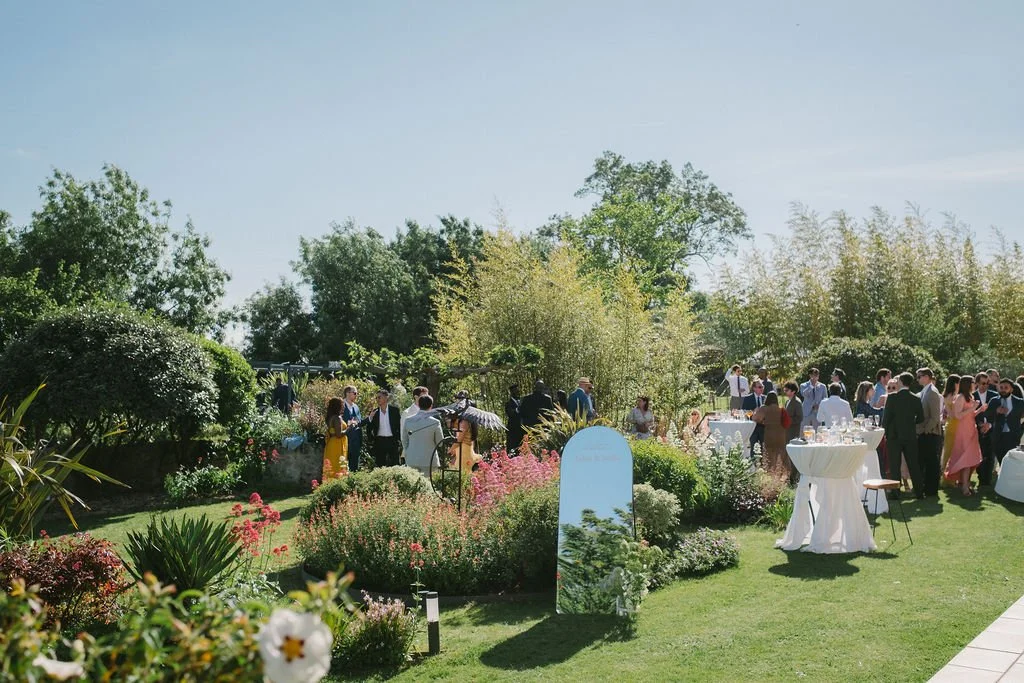 People gathered in a garden for an outdoor event at Le Domaine de Loustalviel on a sunny day, with tables and floral decorations.