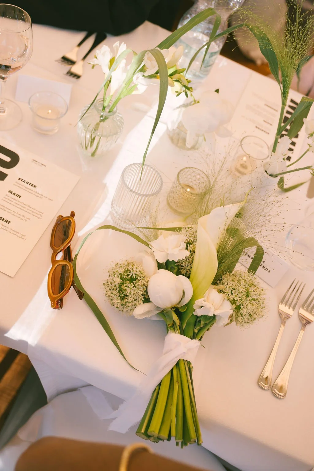 Elegant table setting with a bouquet of white flowers, glasses, candles, and a pair of orange sunglasses on a white tablecloth at a formal event.