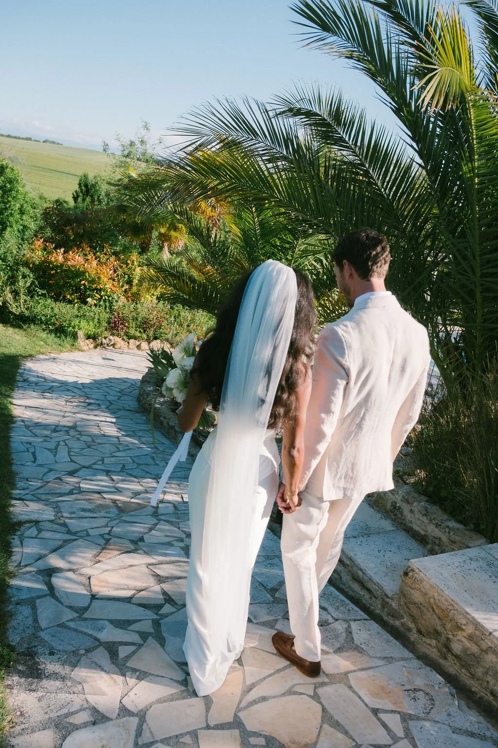 A bride and groom walking hand in hand outdoors on a stone pathway at Le Domaine de Loustalviel, surrounded by lush greenery and palm trees.