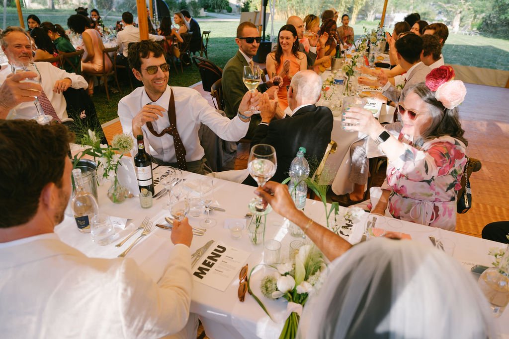 People gathered around a banquet table outdoors celebrating at a wedding, raising glasses for a toast, with greenery in the background.