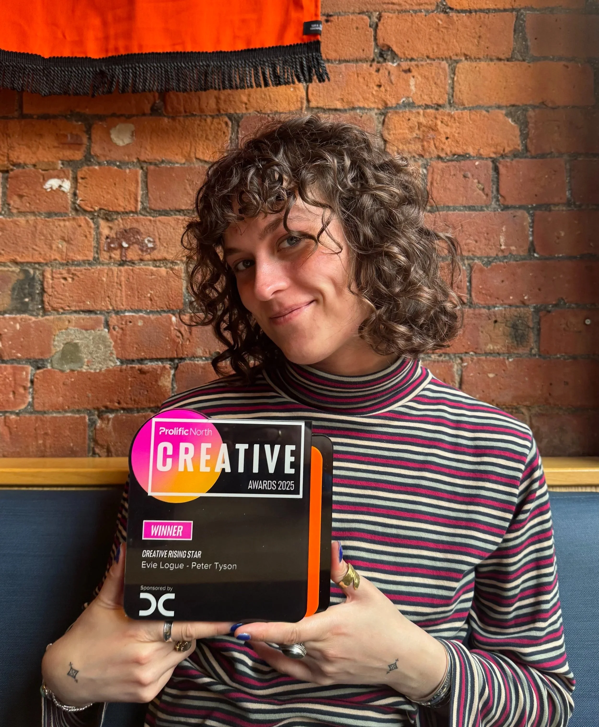 A woman with curly hair and tattoos on her hands holding an award plaque in front of her face, partially obscuring it, against a brick wall background. The plaque reads "Prolific North Creative Awards 2025" and indicates she is a winner.