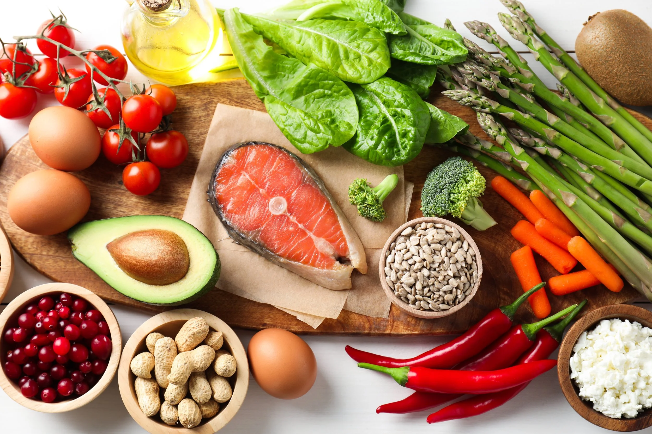 Variety of healthy foods including salmon, leafy greens, asparagus, nuts, eggs, avocados, cherry tomatoes, carrots, hot peppers, broccoli, berries, sunflower seeds, and a bottle of olive oil on a wooden board.