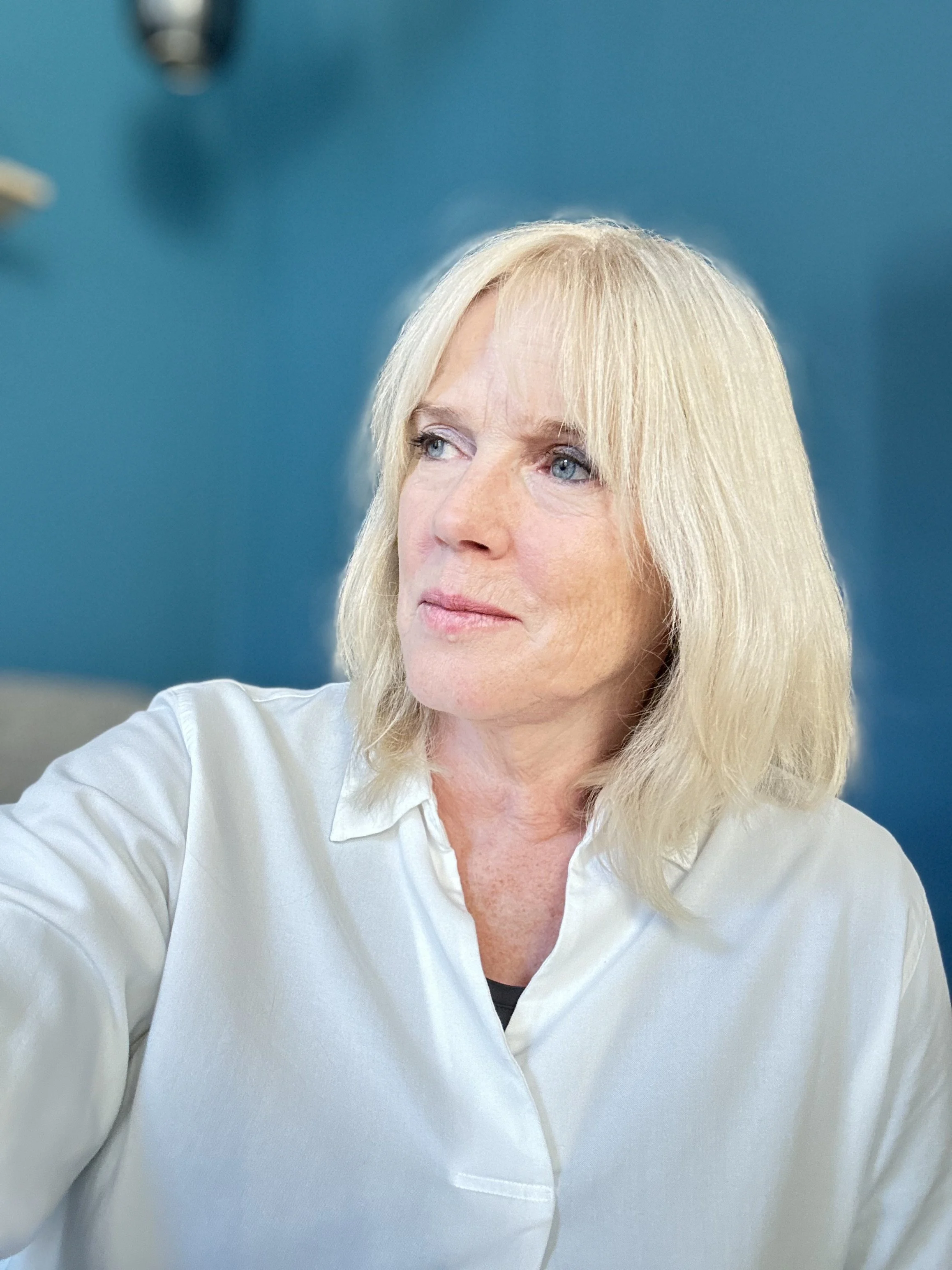 Close-up of a woman with blonde hair and blue eyes wearing a white blouse, looking to her right, against a blue wall background.