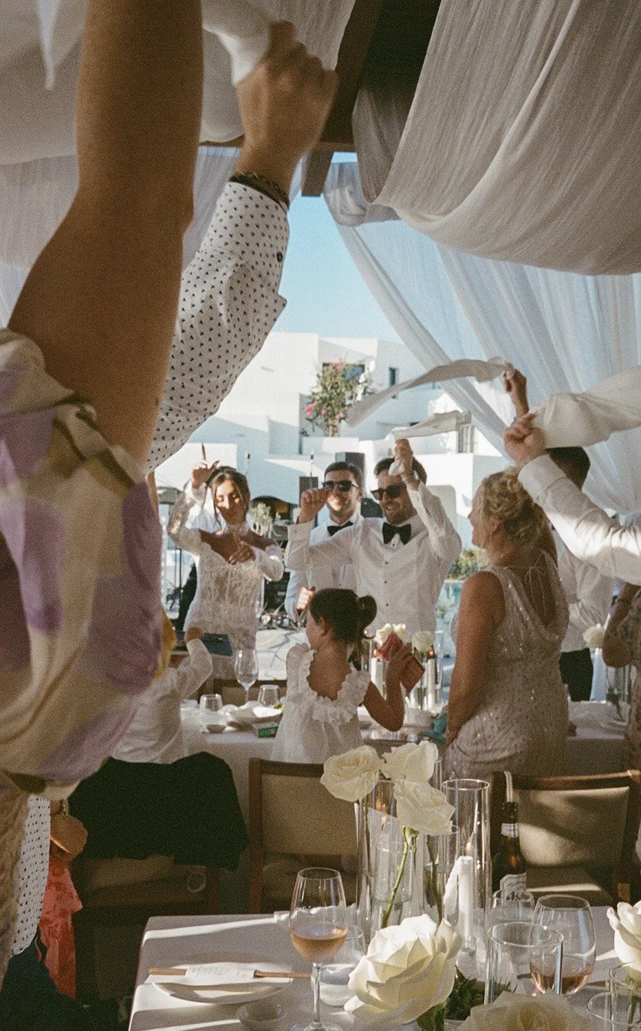 Wedding celebration with guests dancing and waving napkins, decorated tables with flowers and drinks, bright outdoor setting.