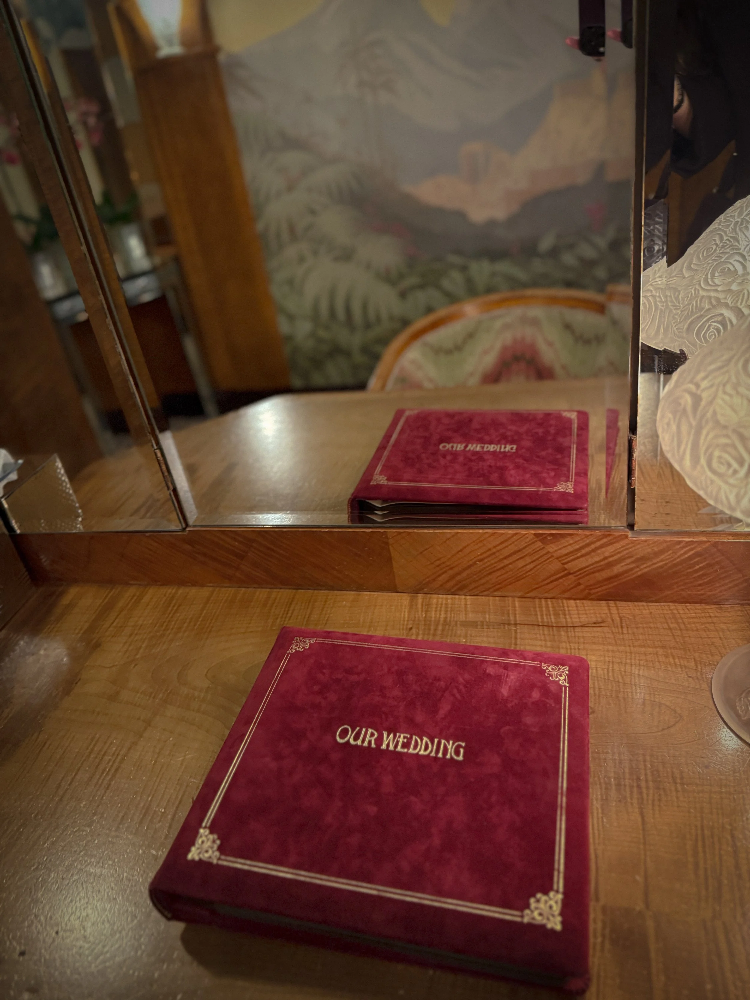 Red velvet vintage wedding album on a wooden table in front of a mirrored vanity with floral wallpaper in the background in London Claridge's toilets.