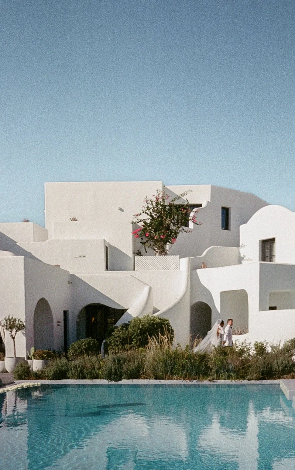 Bride and groom newlywed in front of a white Mediterranean-style building in Santorini with a pool and plants under a clear blue sky.