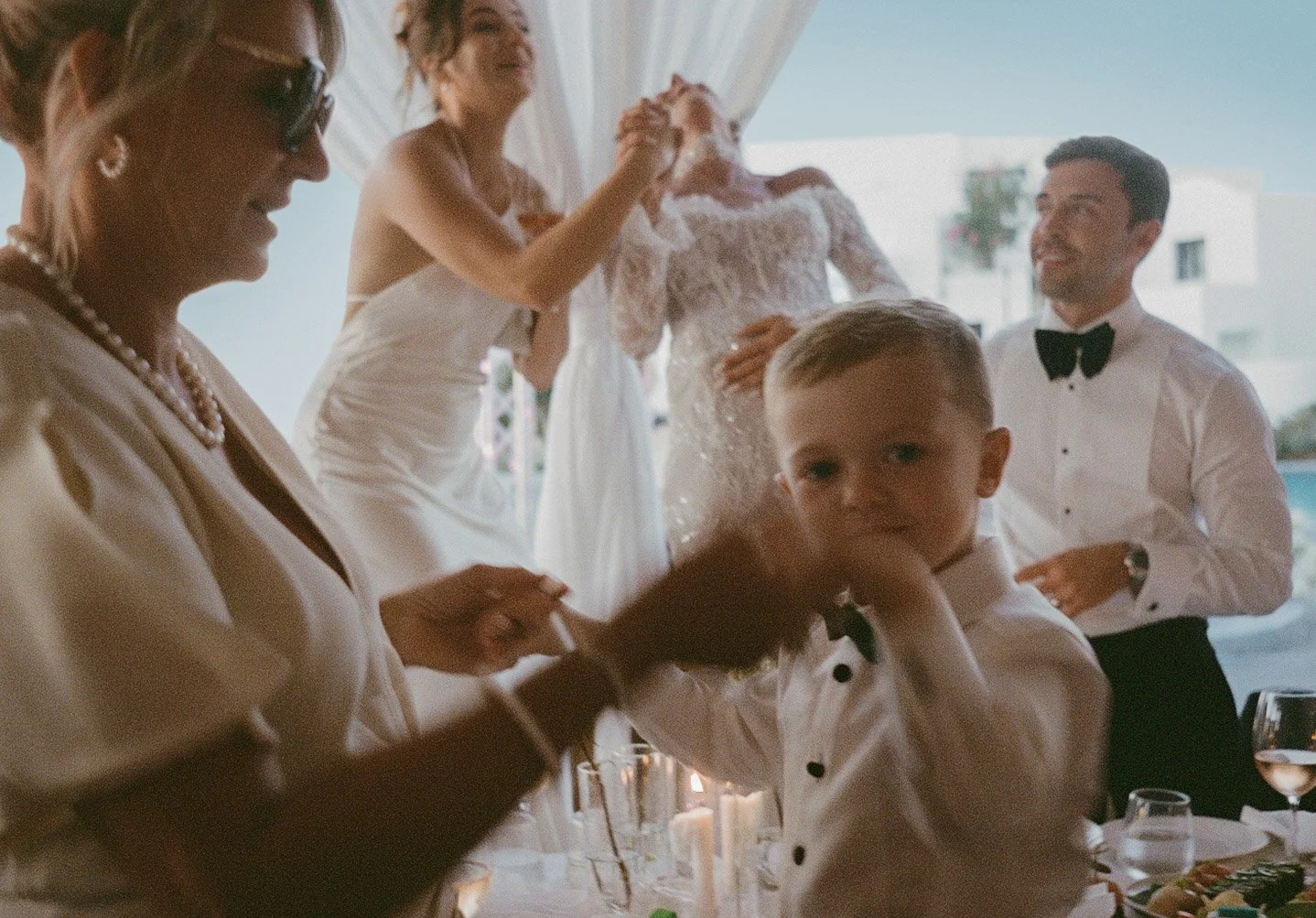 Group of people celebrating indoors at a Santorini wedding, including a child in a tuxedo and adults in formal attire.