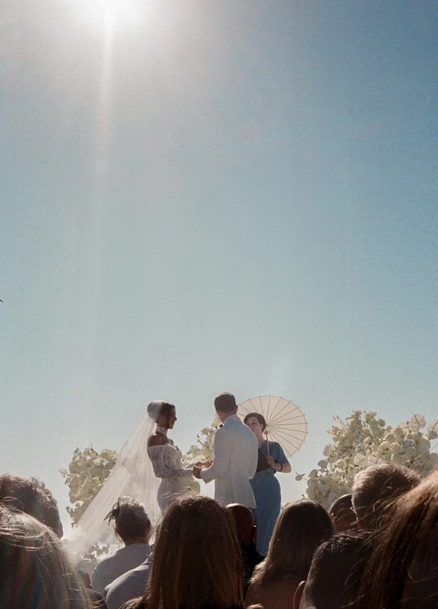 Outdoor wedding ceremony with bride and groom, officiant holding an umbrella, clear sky, and floral decorations.