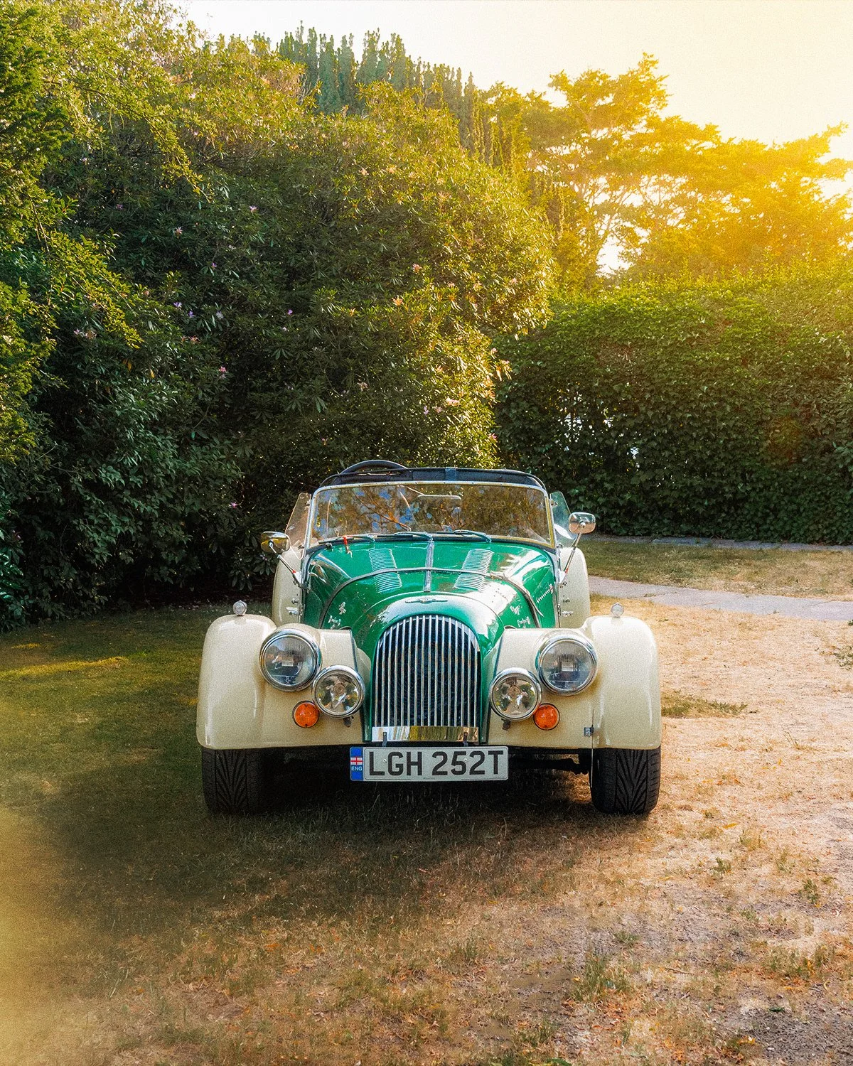 A vintage green and cream-colored convertible car parked on a grassy patch with trees and bushes in the background, illuminated by sunlight.