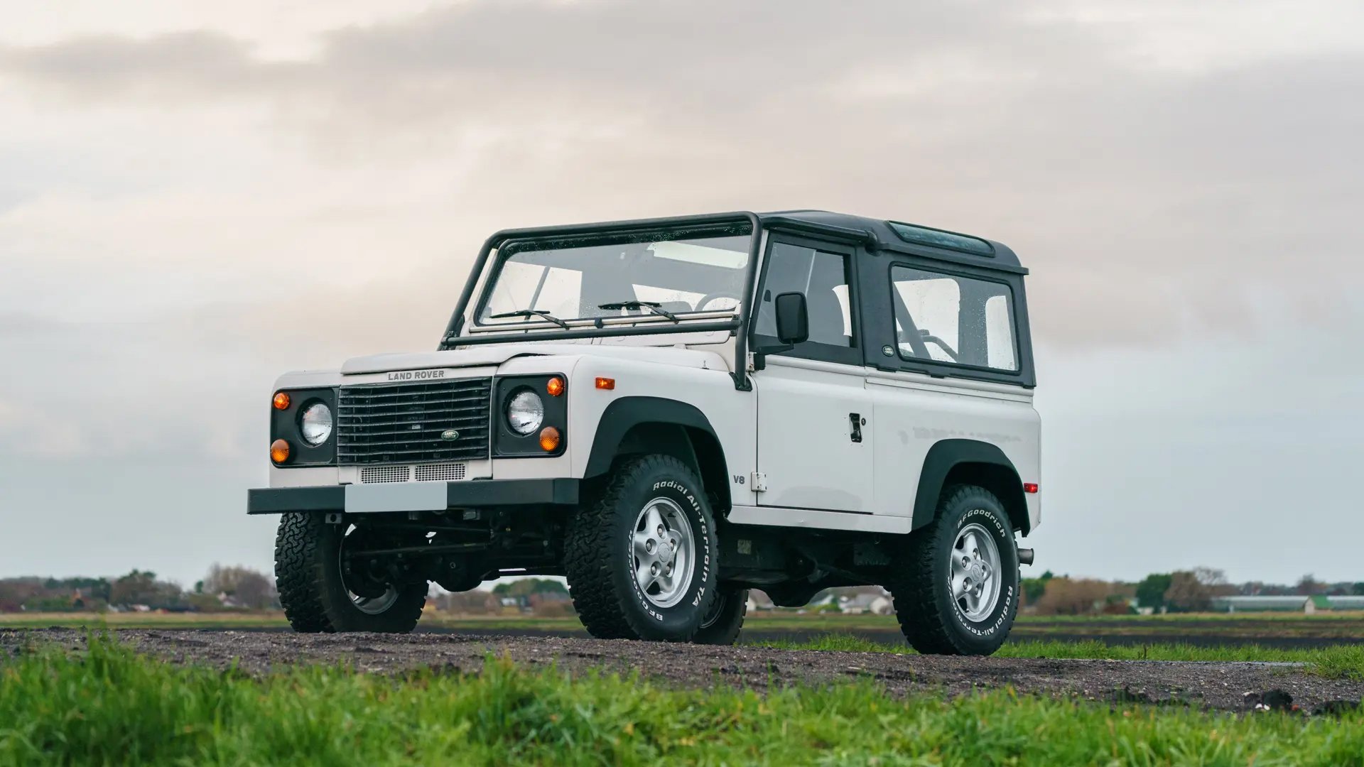 A white Land Rover Defender parked on a grassy area with a cloudy sky in the background.