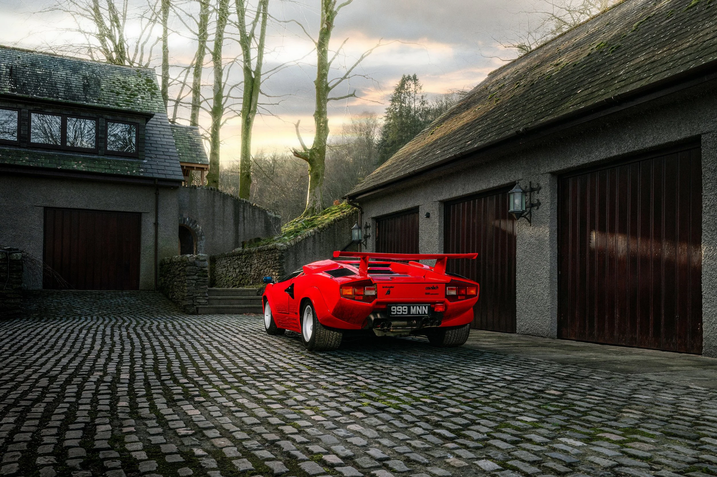 A red Lamborghini Countach sports car parked on a cobblestone driveway between two garage buildings at sunset.