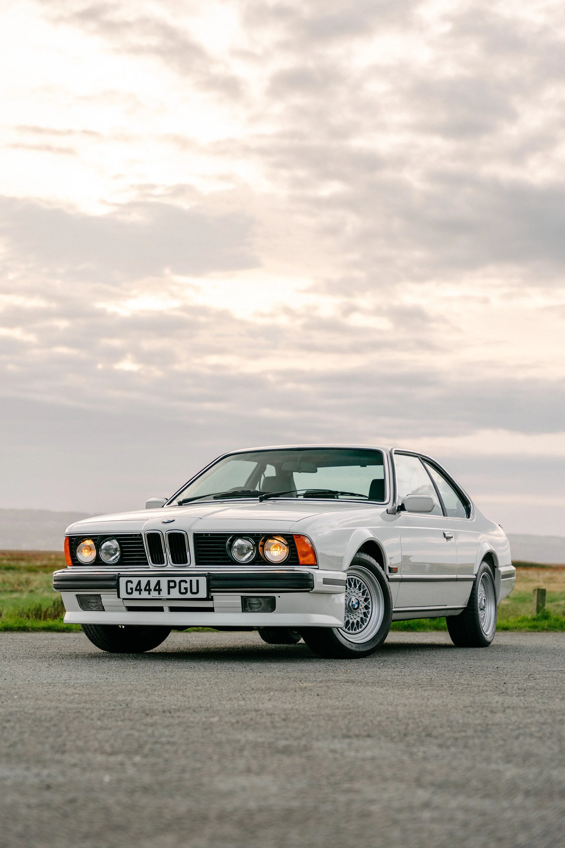 A classic white BMW E24 6 Series coupe parked on a paved surface with a cloudy sky and open landscape in the background.