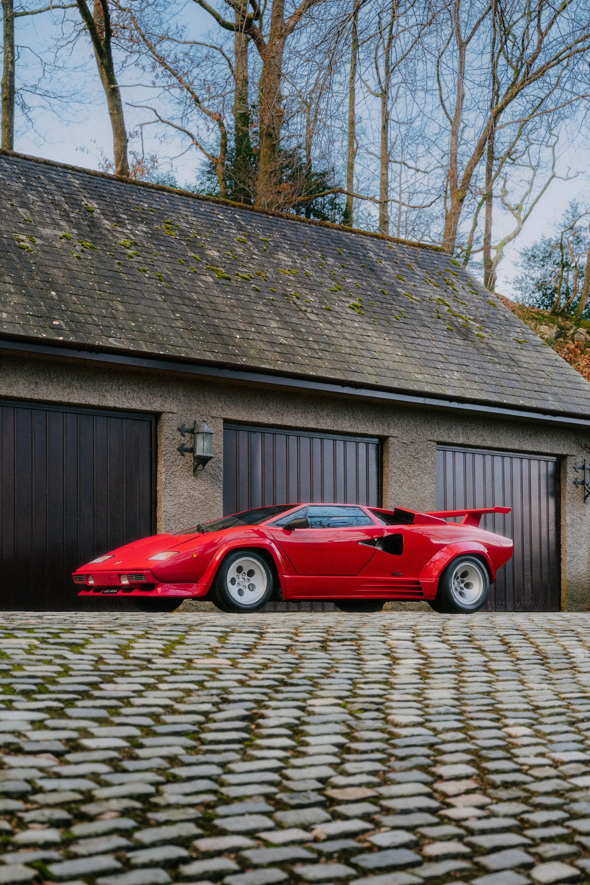 A classic red Lamborghini Countach parked on a cobblestone driveway in front of a gray garage with black doors. The house has a sloped roof with moss and is surrounded by trees with bare branches.