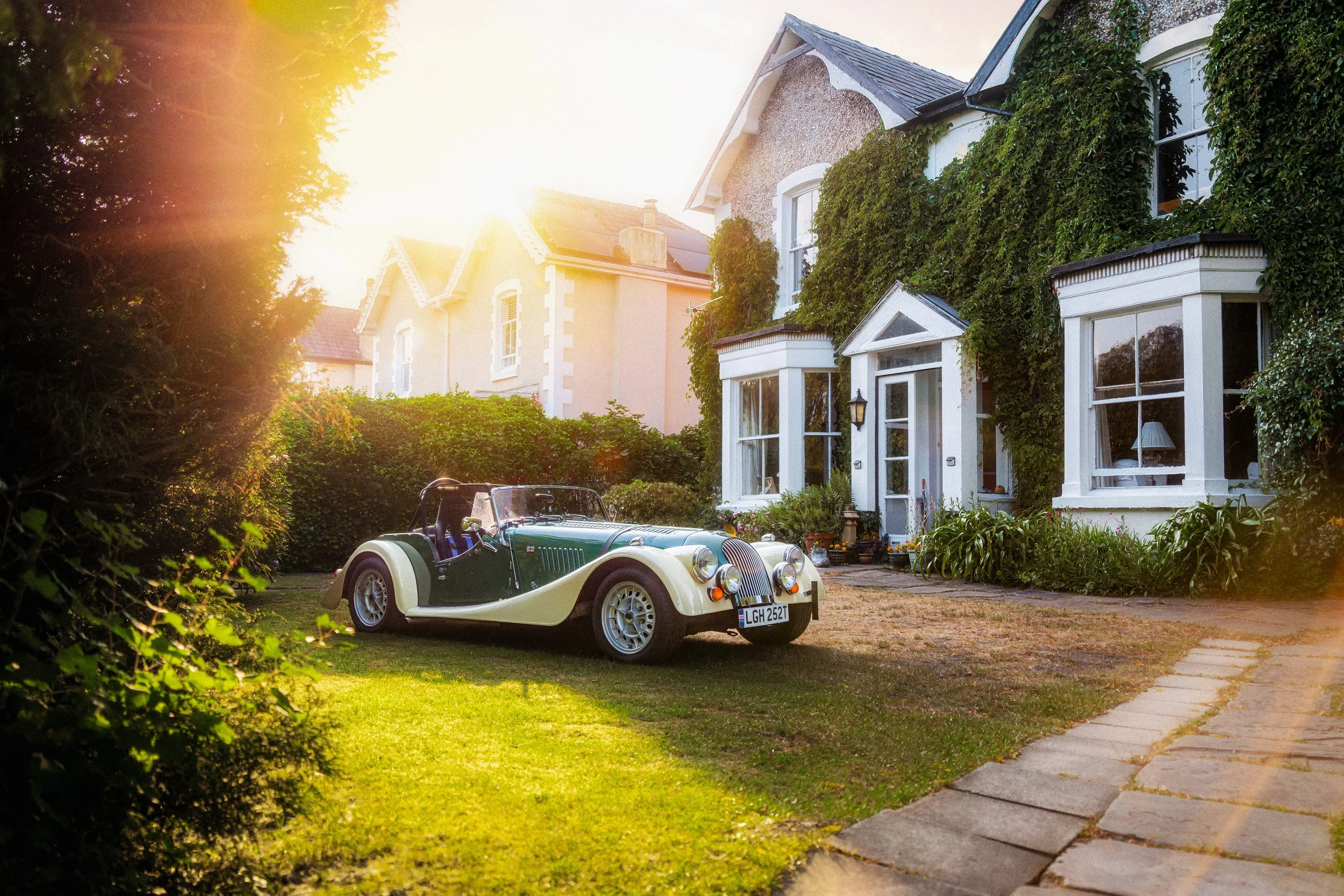 A vintage green and cream-colored convertible car parked on a lawn in front of a white house with ivy-covered walls. The house has large bay windows, a small front porch, and a garden with plants and flowers. The scene is lit by bright sunlight, crea