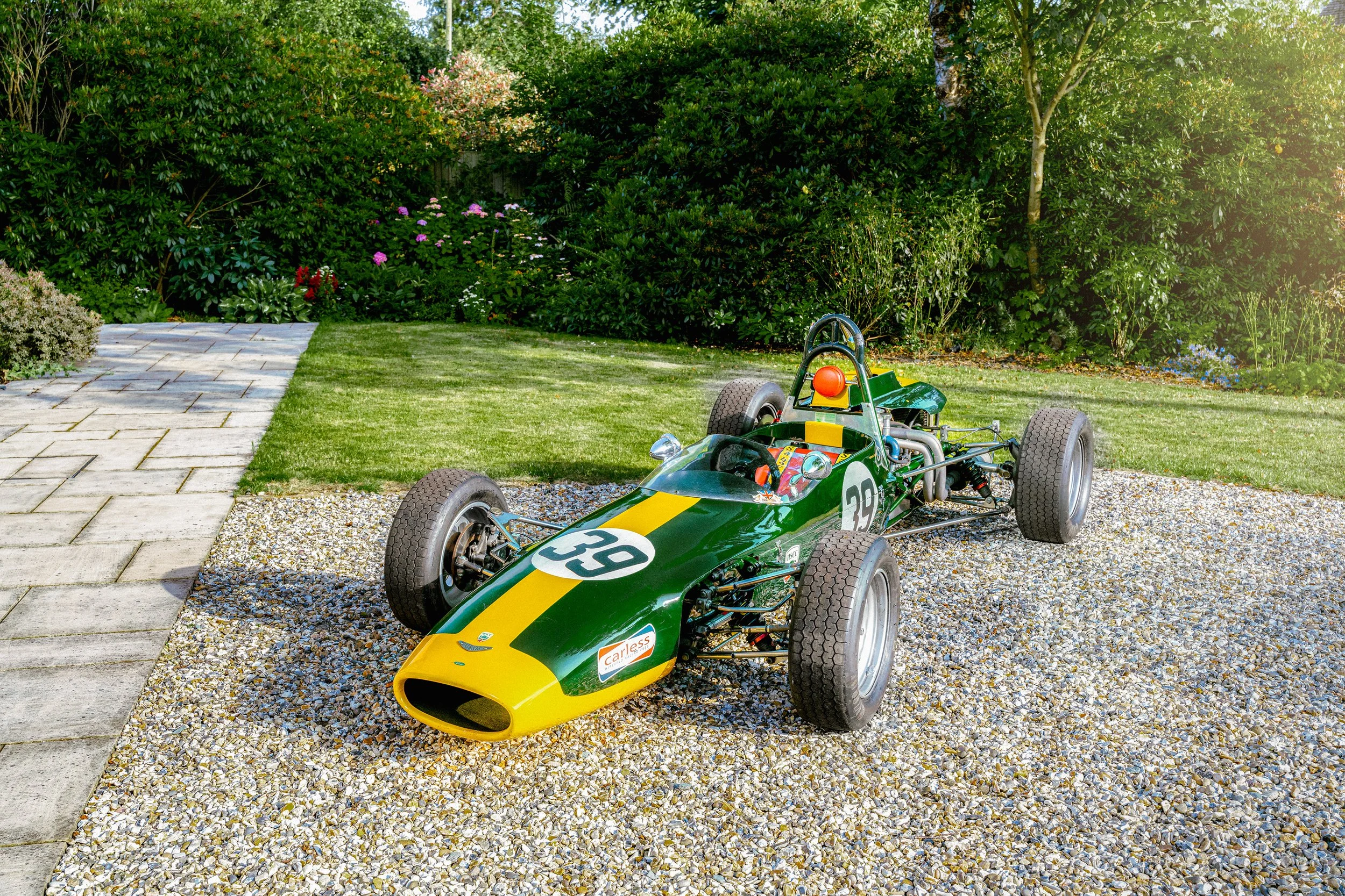 A vintage green and yellow race car with the number 39 on its nose, parked on a gravel surface in a garden with green bushes and trees.