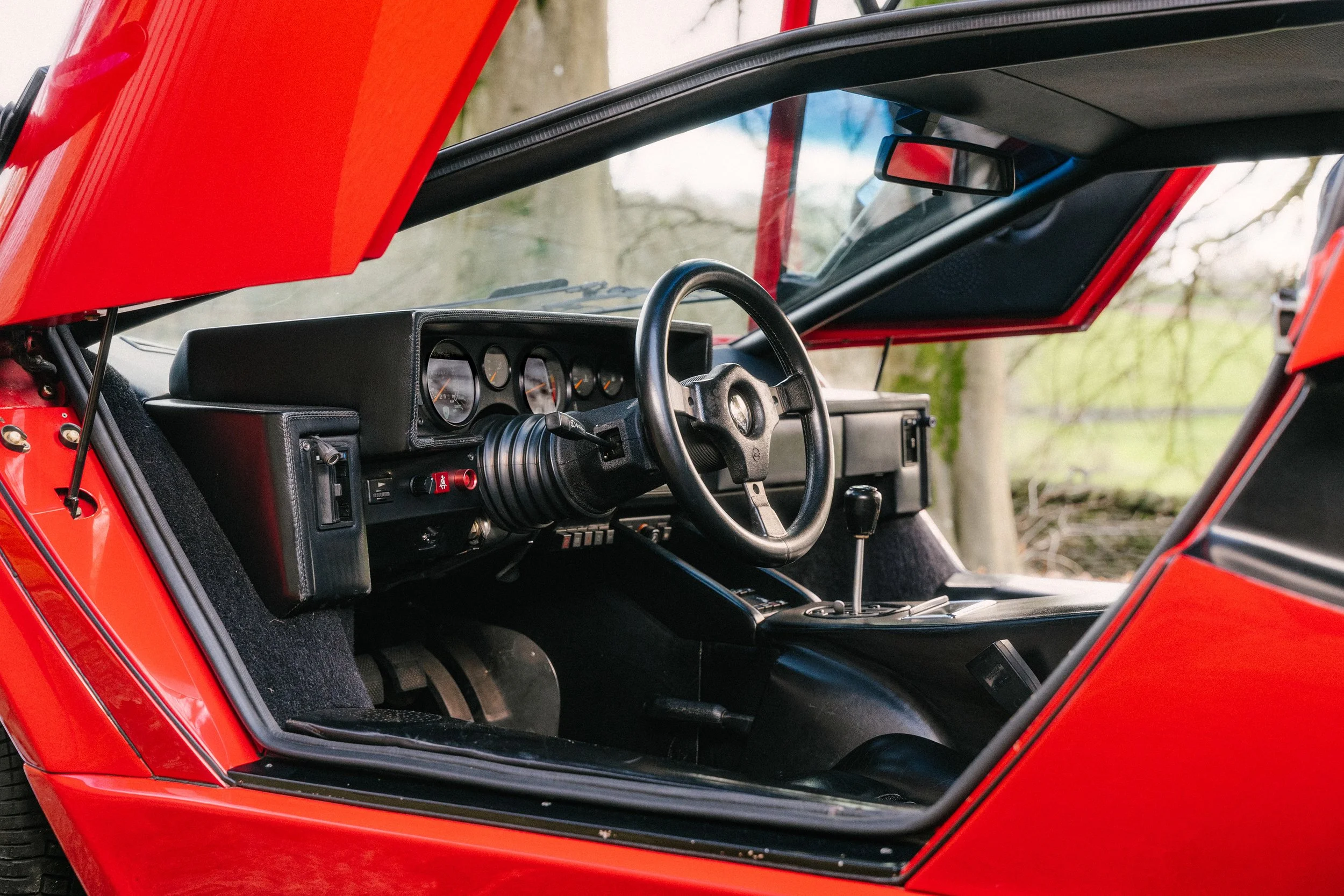 Interior of a red sports car, showing the dashboard with analog gauges, a steering wheel, and gear shift, with the driver's side door open and a blurred outdoor background.